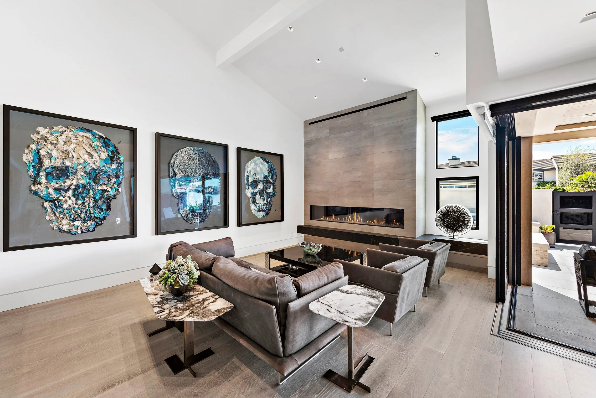 Modern living room with three framed artwork skull posters on white wall, gray sofa and armchairs, a marble side table, and a fireplace, with large windows showing outdoor patio and neighborhood.