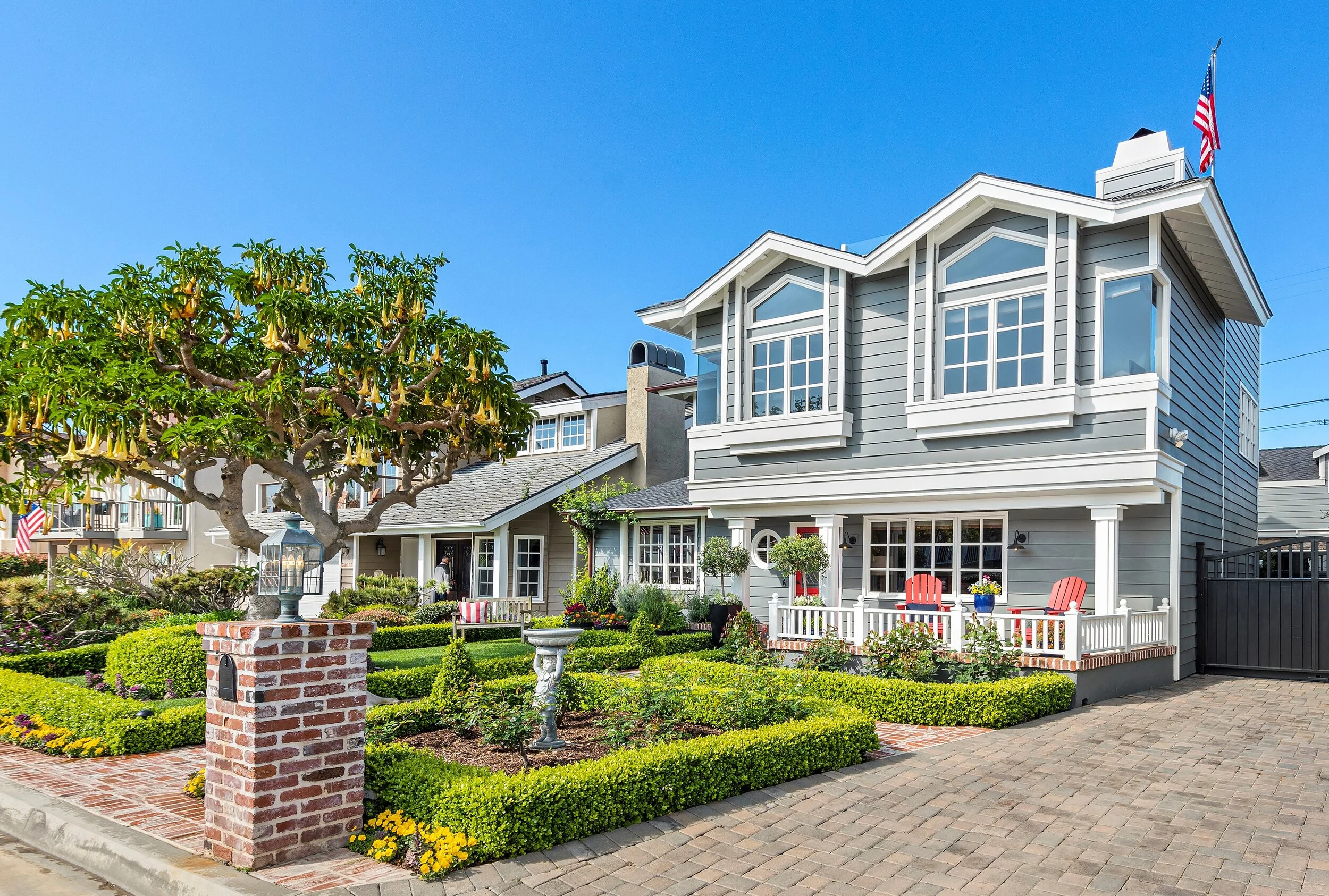A charming two-story gray house with white trim, a landscaped front yard with neatly trimmed hedges, yellow flowers, and garden ornaments, a brick mailbox, and a driveway, under a bright blue sky.