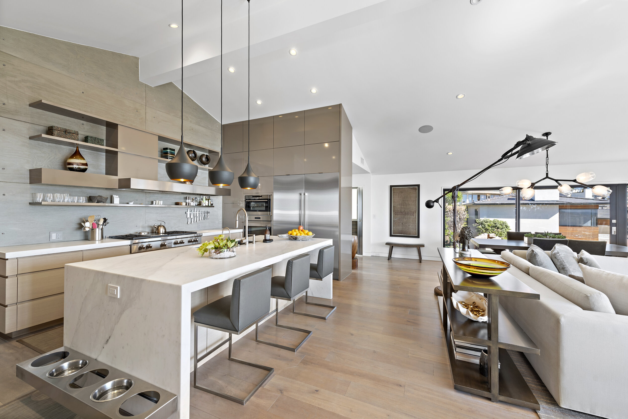 Modern open-concept kitchen and living room with a large white island, gray bar stools, stainless steel appliances, pendant lights, and a cozy seating area with a white sofa and a large window overlooking greenery.