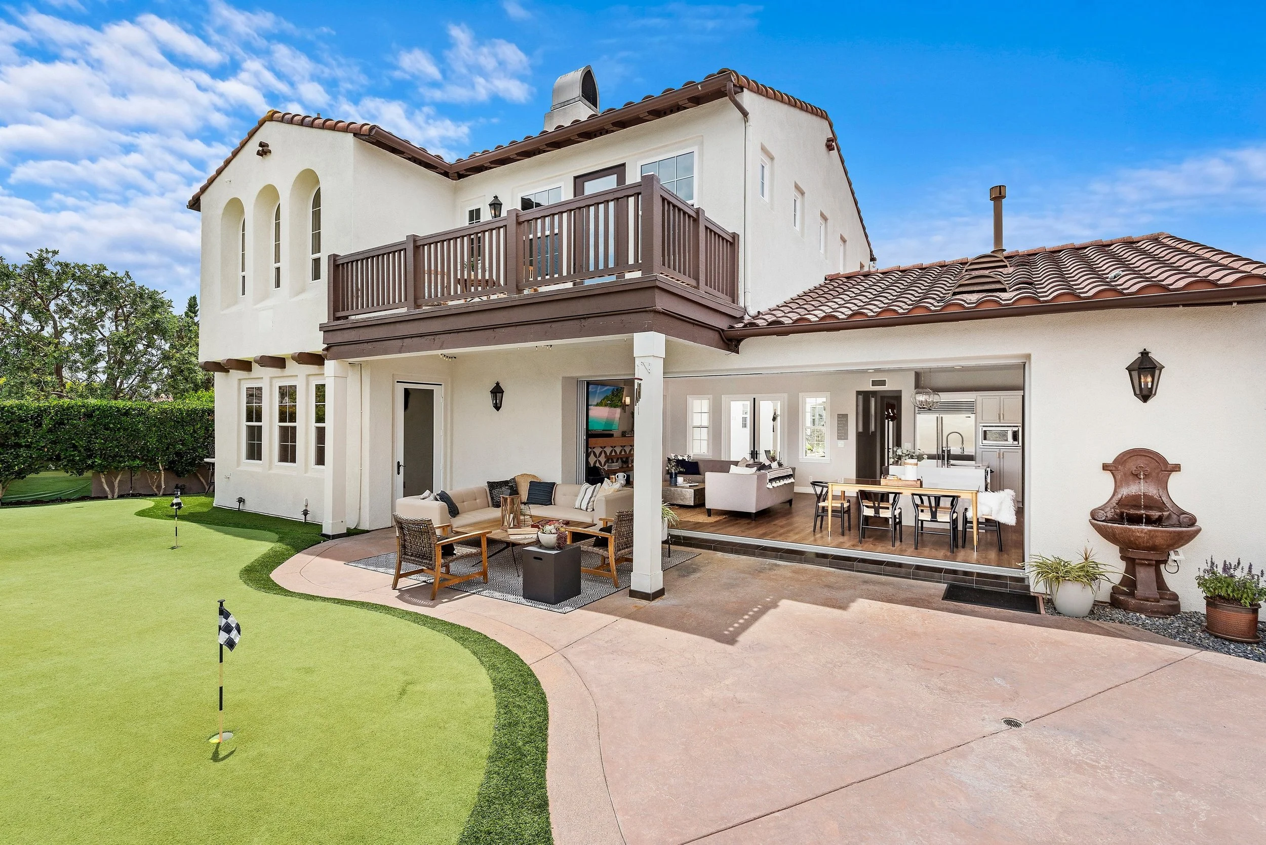 Backyard view of a two-story house with a covered patio, outdoor seating area, a well-maintained lawn, and a putting green, under a blue sky with scattered clouds.