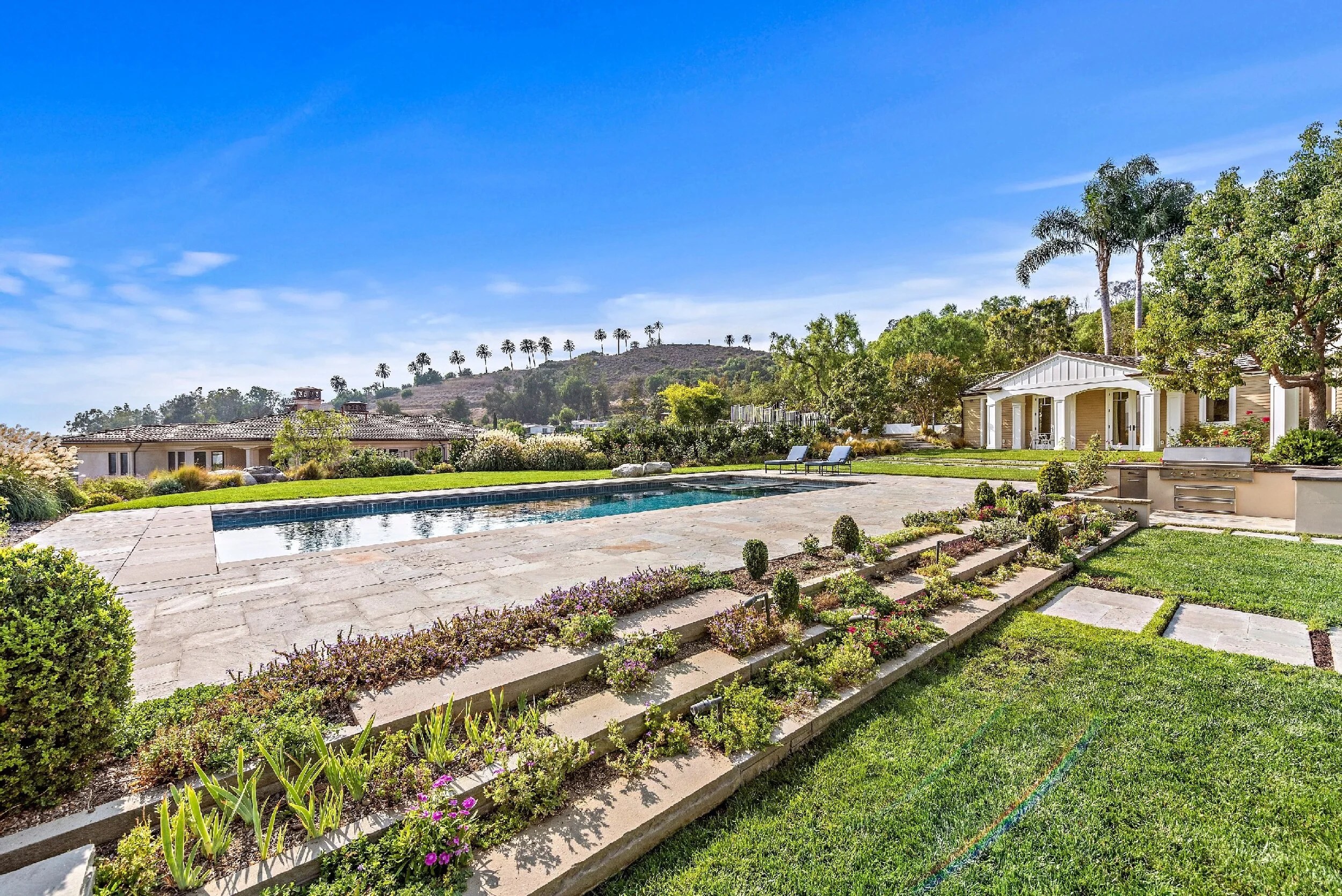 Luxurious backyard with a swimming pool, stone patio, lush green lawn, and scenic hill with trees in the background under a bright blue sky.