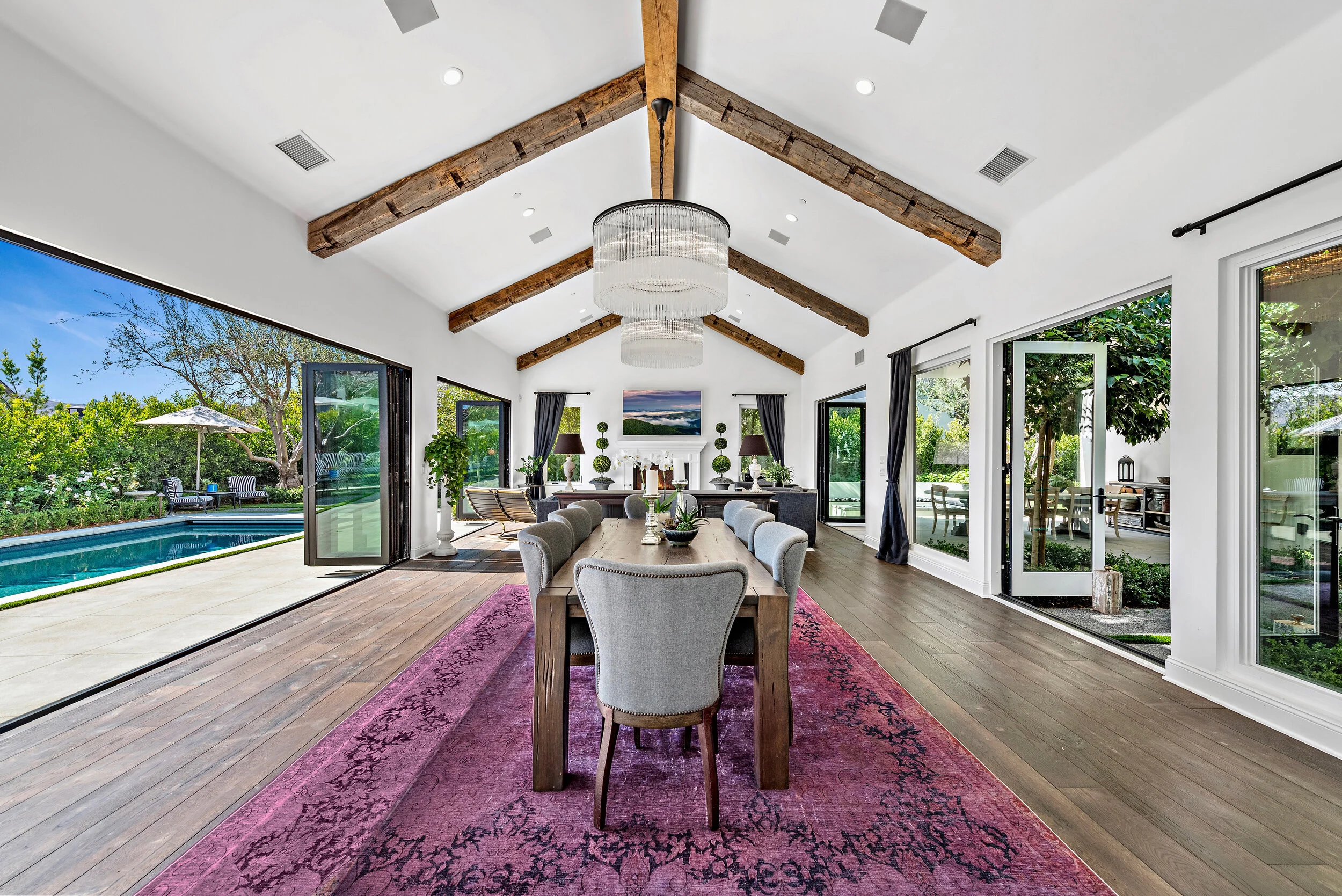 Contemporary dining room with large windows and sliding glass doors opening to backyard with pool, wooden beams on white vaulted ceiling, chandelier, and modern furniture.