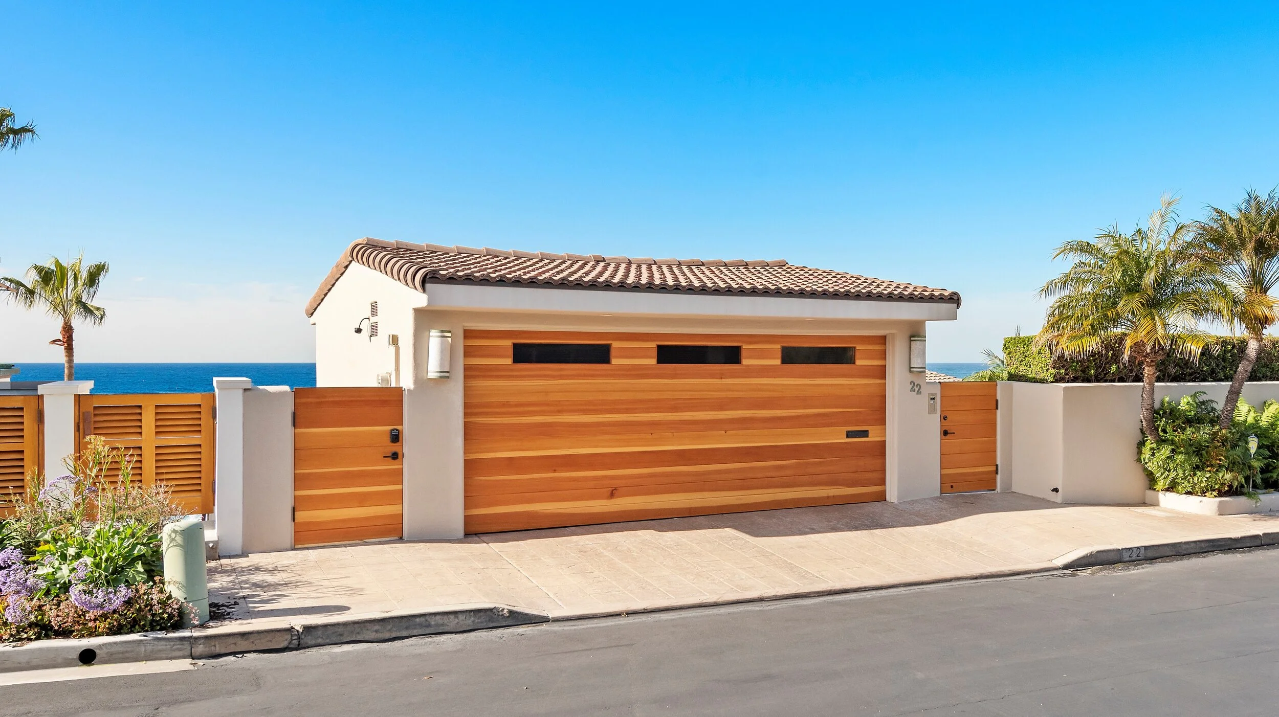 Modern house with a wooden garage door, palm trees, and ocean in the background under a clear blue sky.