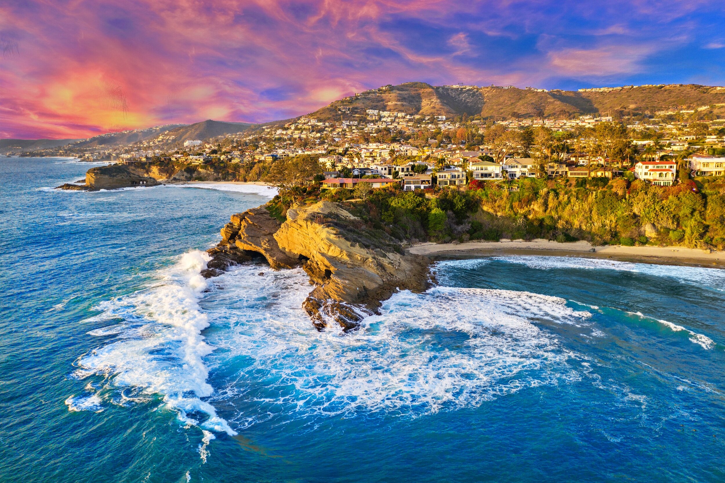 A coastal landscape during sunset with waves hitting rocks, a sandy beach, and a hillside covered with houses and greenery, under a colorful sky.