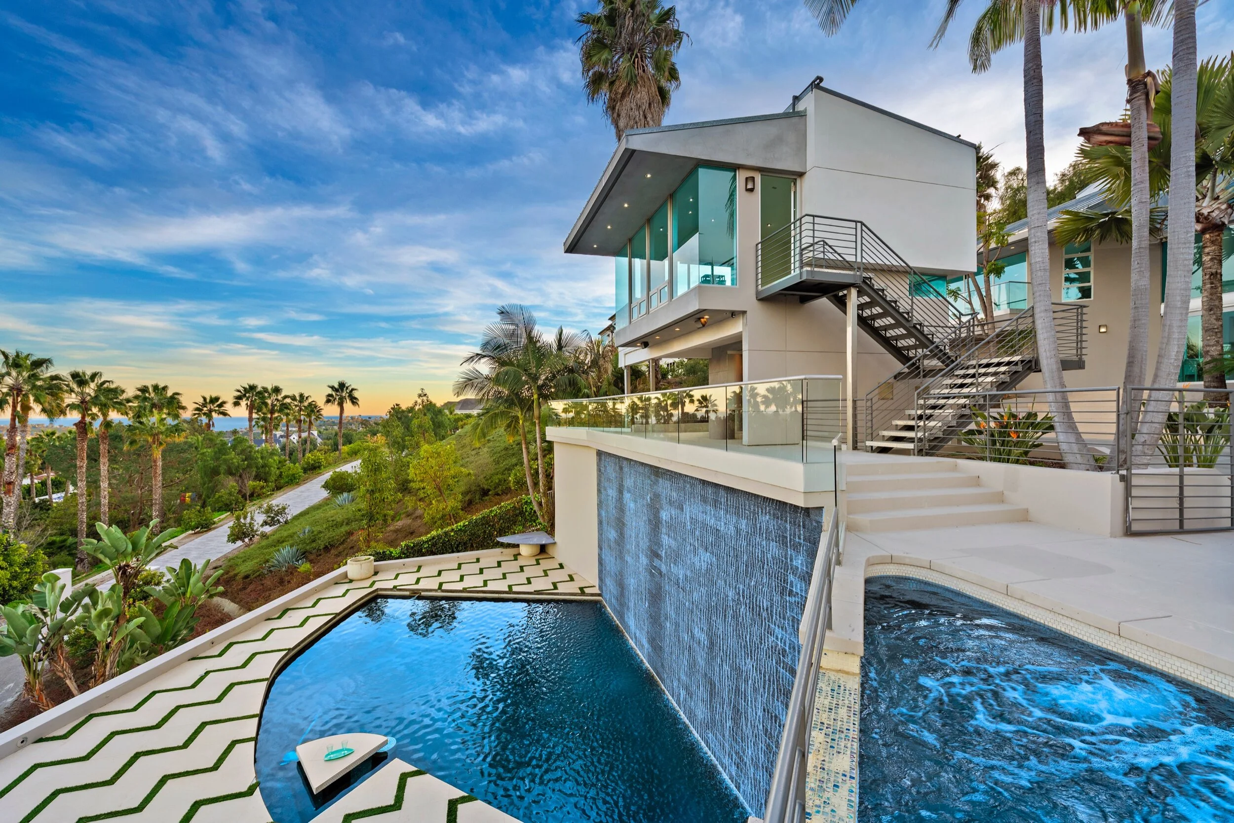 Modern house with outdoor pool, waterfall feature, and hillside view with palm trees at sunset.