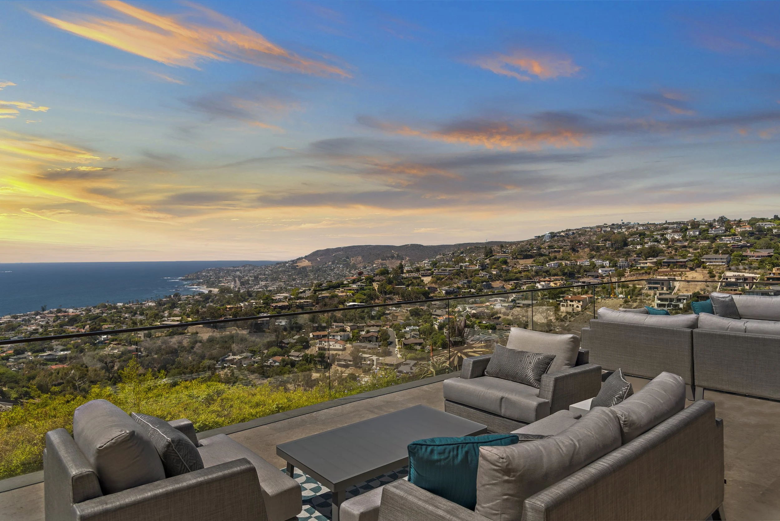 Outdoor patio with gray couches and chairs, overlooking a hilly landscape and ocean at sunset, with a colorful sky and clouds.