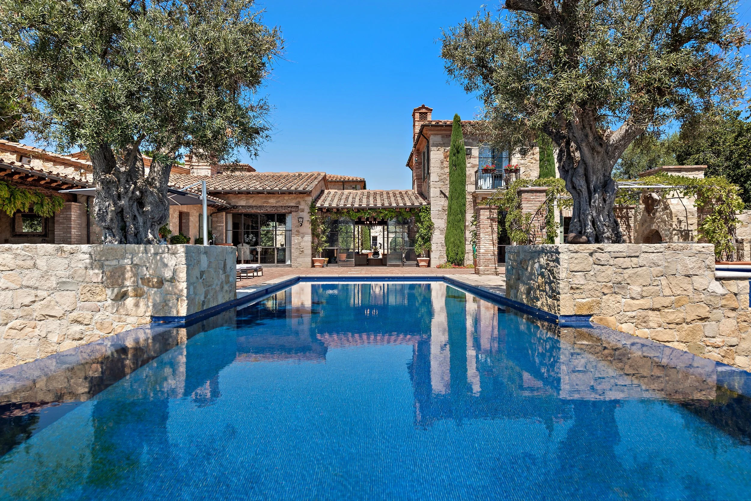 A luxurious outdoor swimming pool in front of a stone house with a terracotta roof, surrounded by trees and greenery, under a clear blue sky.