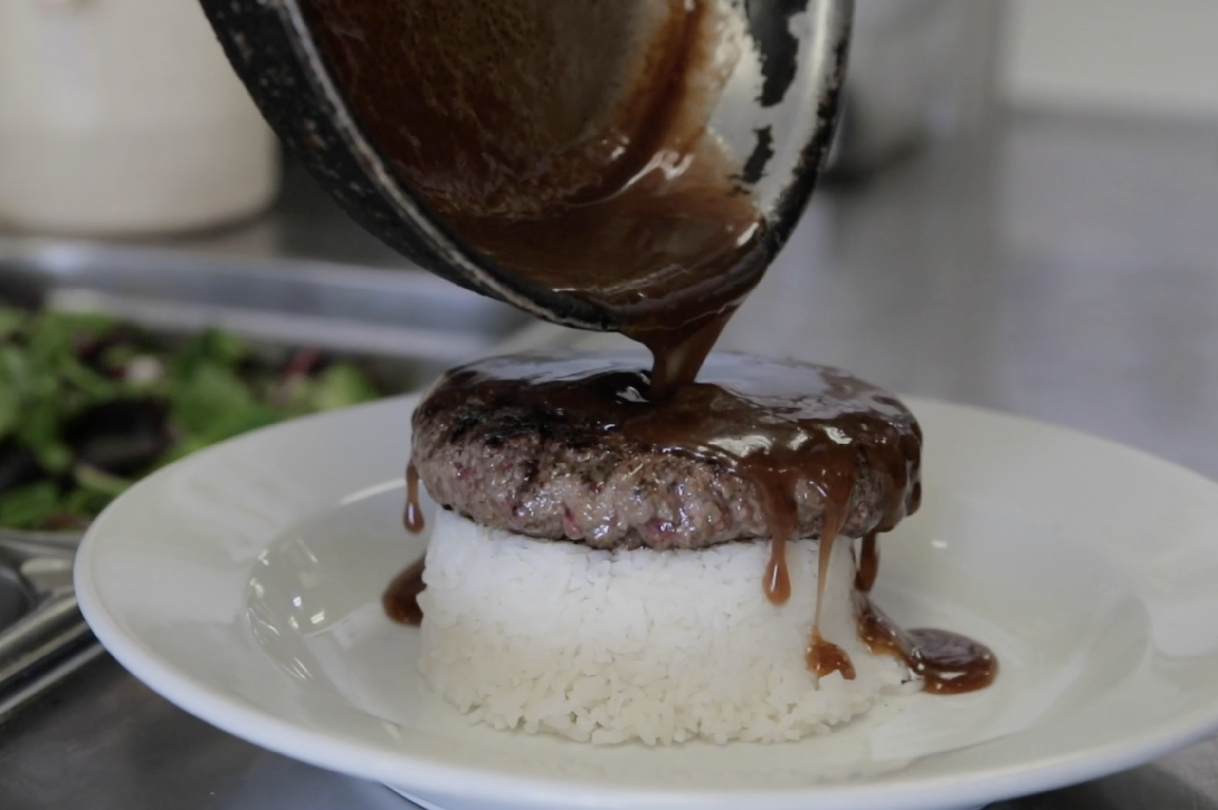 Ground beef patty with gravy being poured over a bed of rice on a white plate.