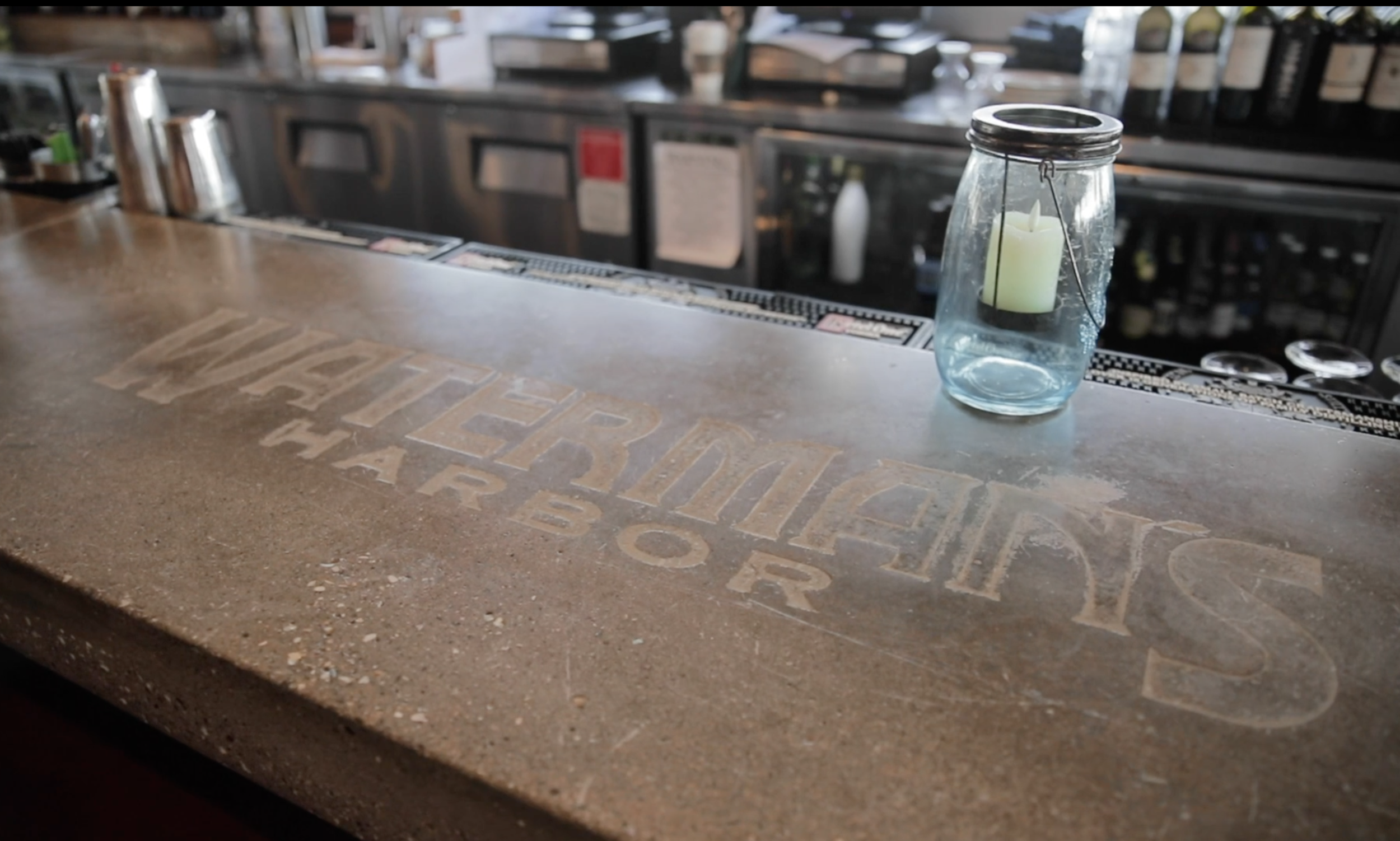 Bar counter with a glass jar holding a green candle, and a faded logo on the surface that reads "Patterson's Bar" in a bar or restaurant setting.