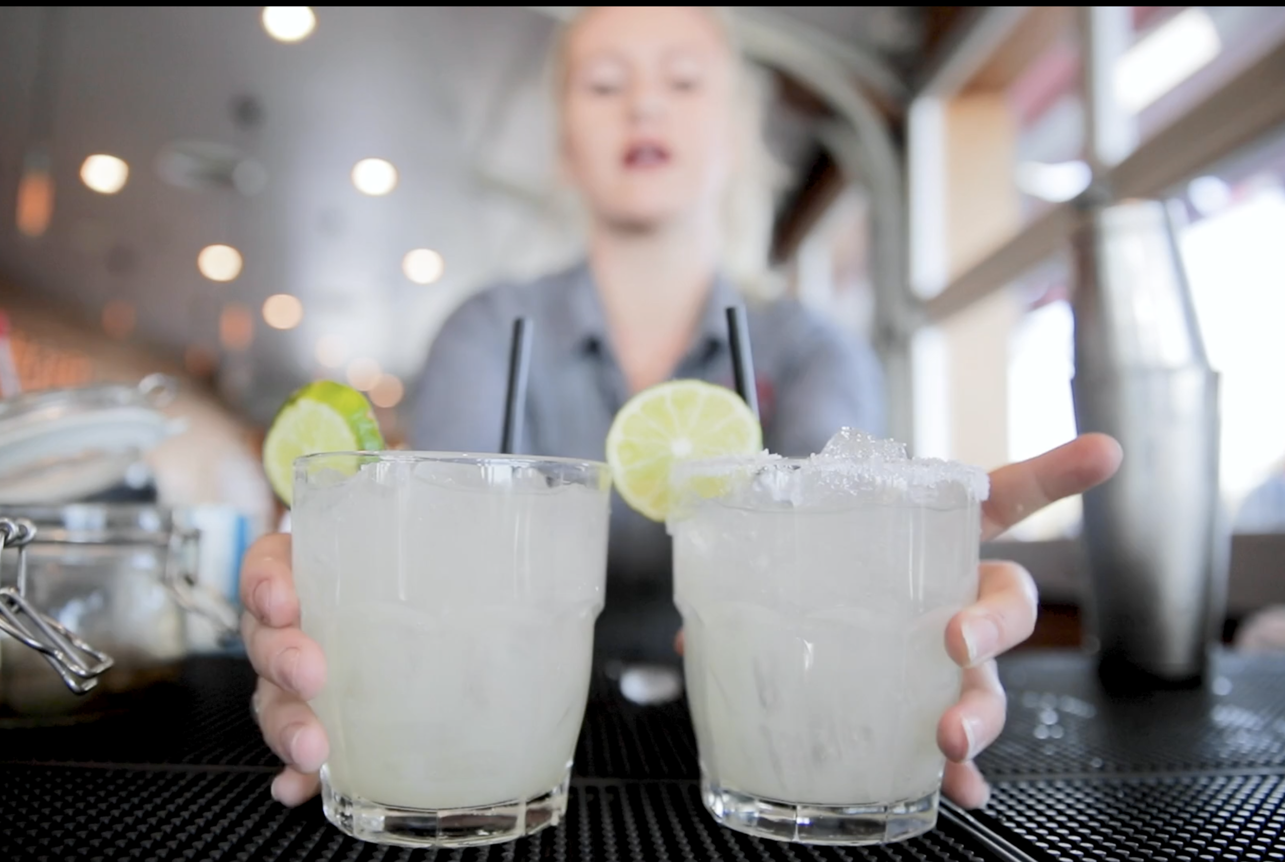 A bartender presents two cocktail glasses garnished with lime slices on a bar counter in a restaurant or bar setting.