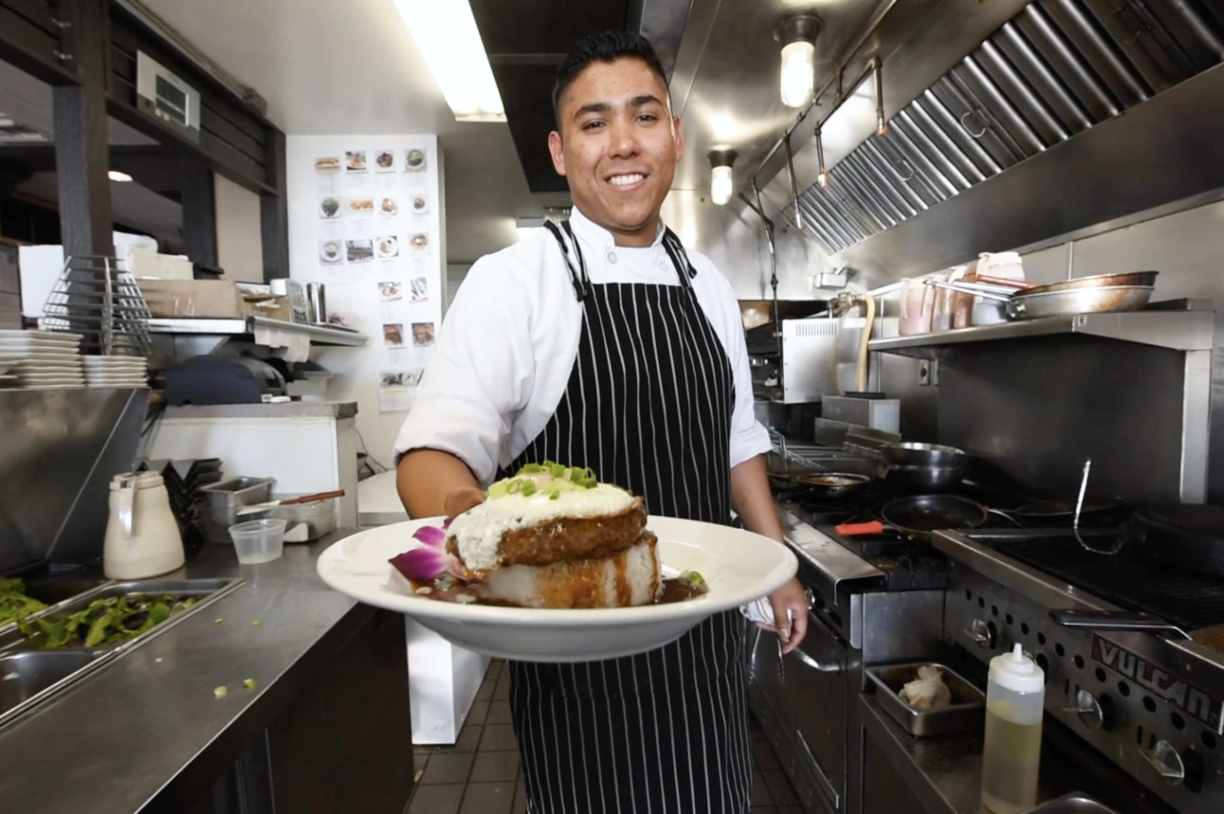 A chef in a striped apron and white shirt smiling and holding a plate of food in a restaurant kitchen.
