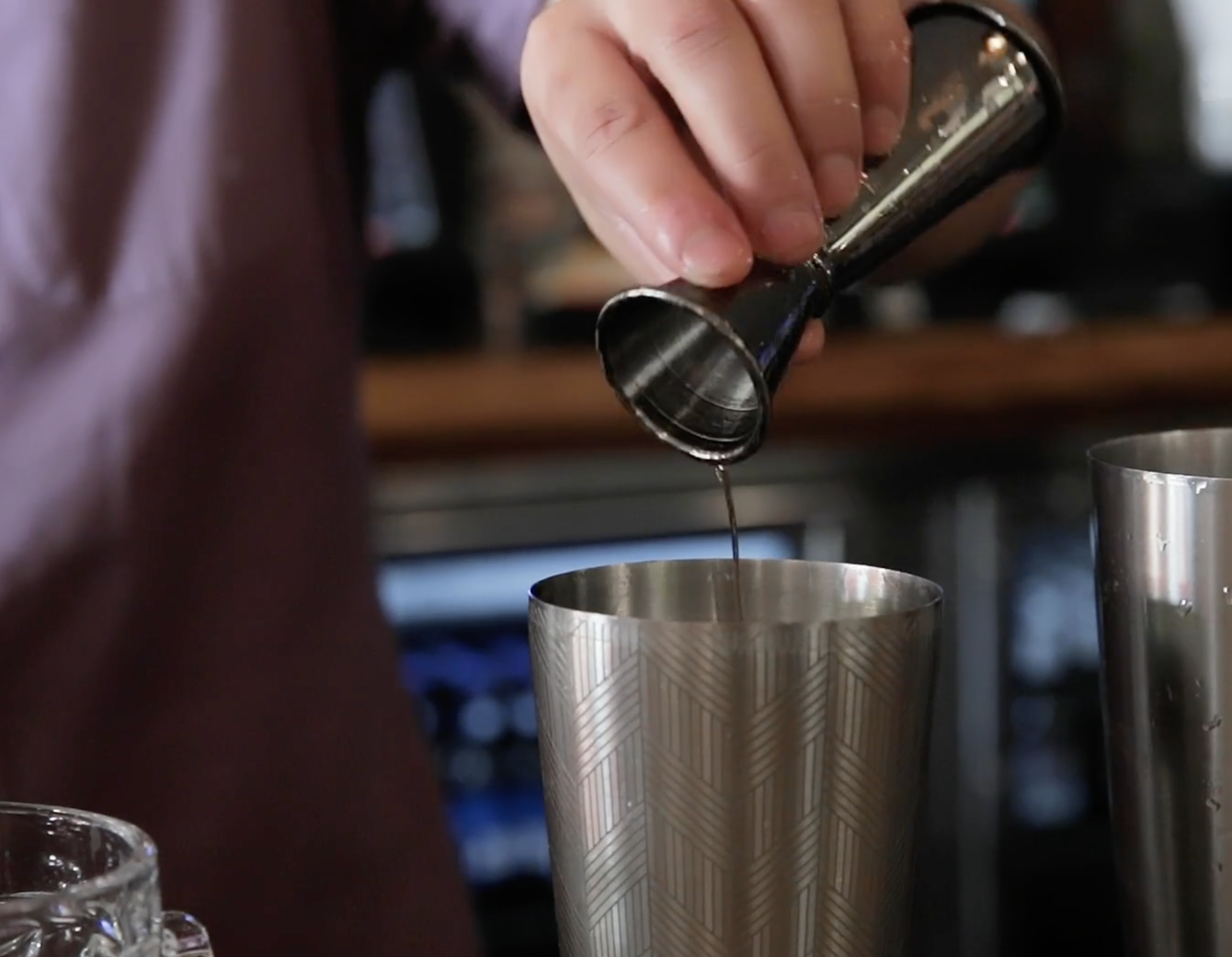 Someone pouring liquid from a small metal cocktail shaker into a tall, patterned metal cup.