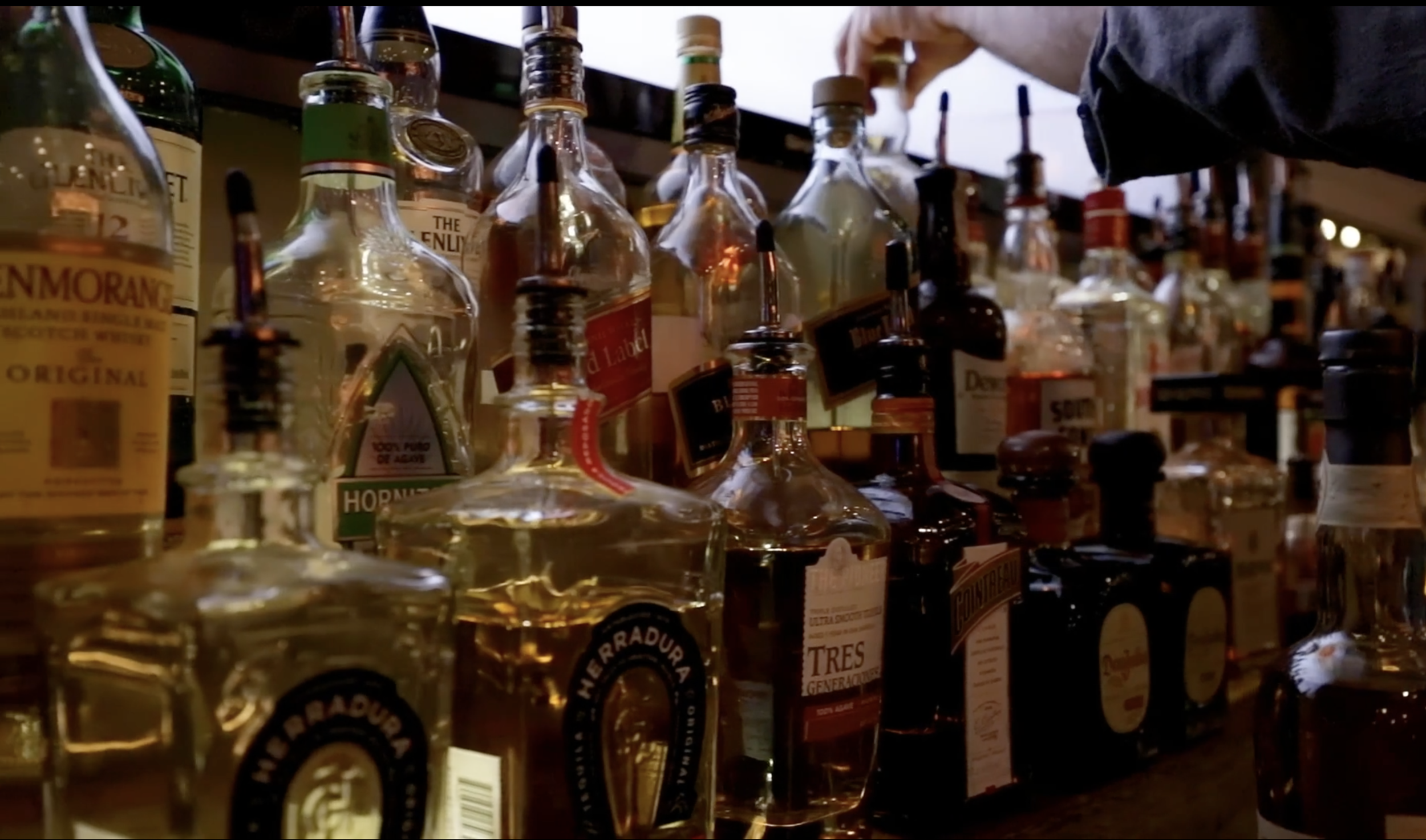 Assorted bottles of whiskey on a bar shelf, with a person reaching for a bottle in the background.