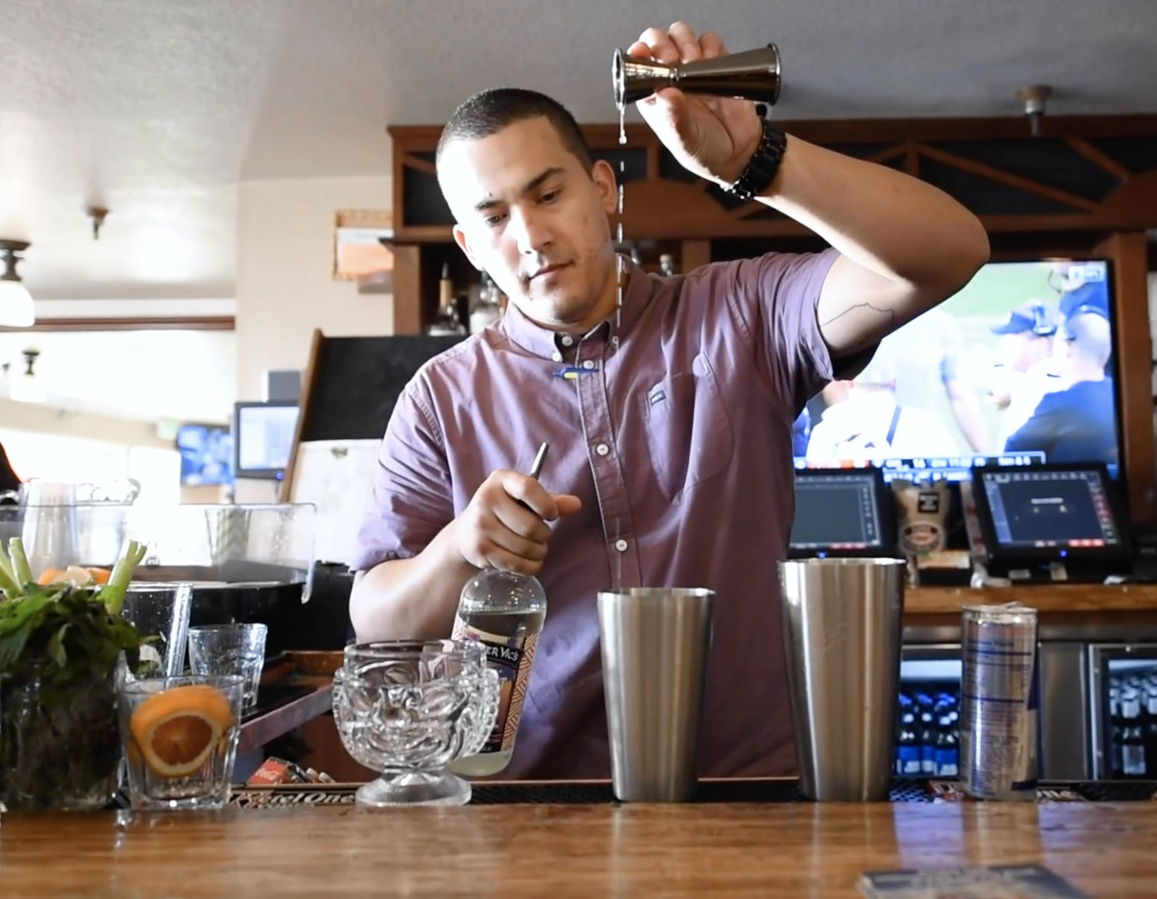 A bartender pouring a clear liquid into a cocktail shaker at a bar counter, with various bar tools and glasses, and a large television screen displaying sports in the background.