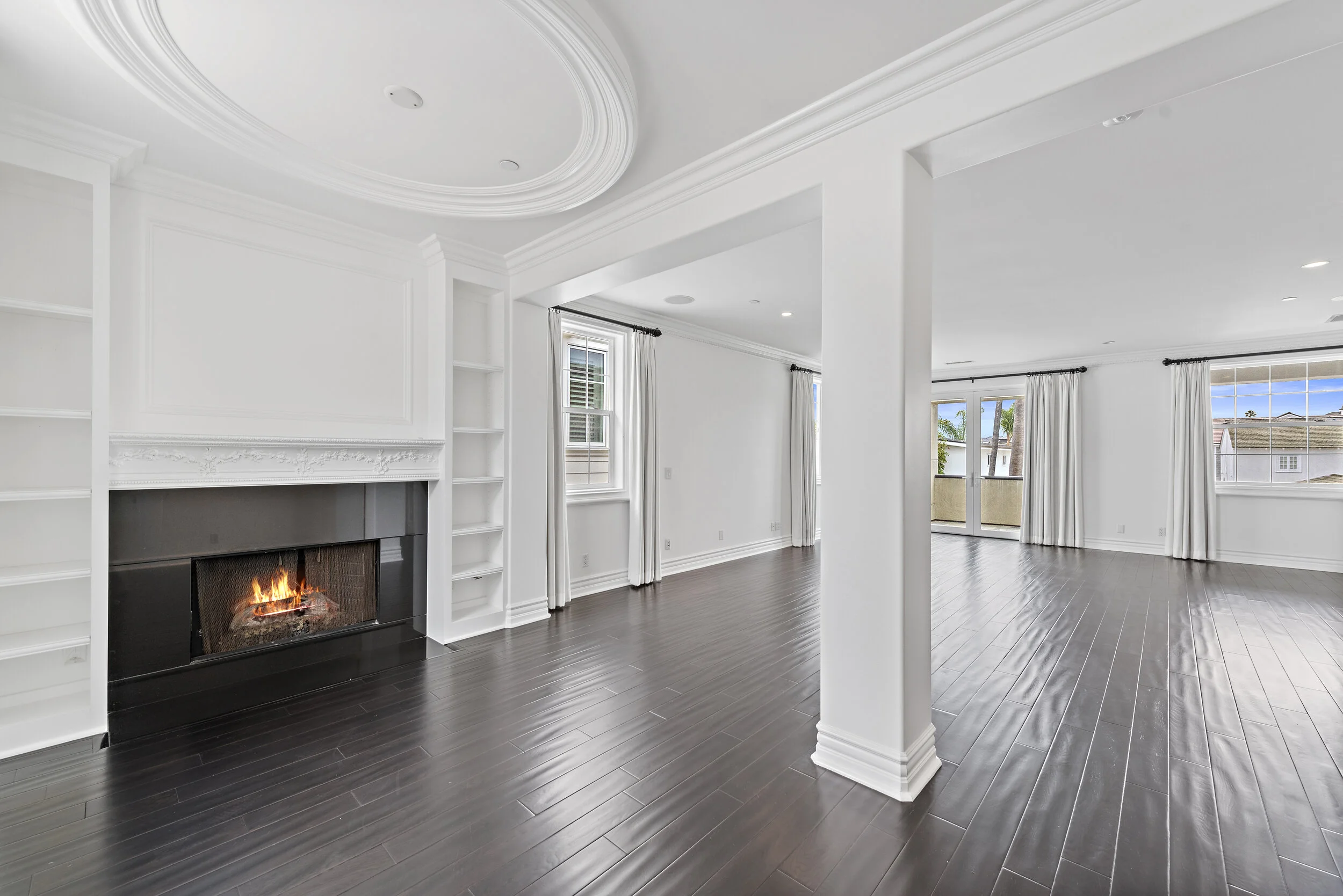Empty living room with white walls, dark wood flooring, fireplace, and large windows with white curtains.