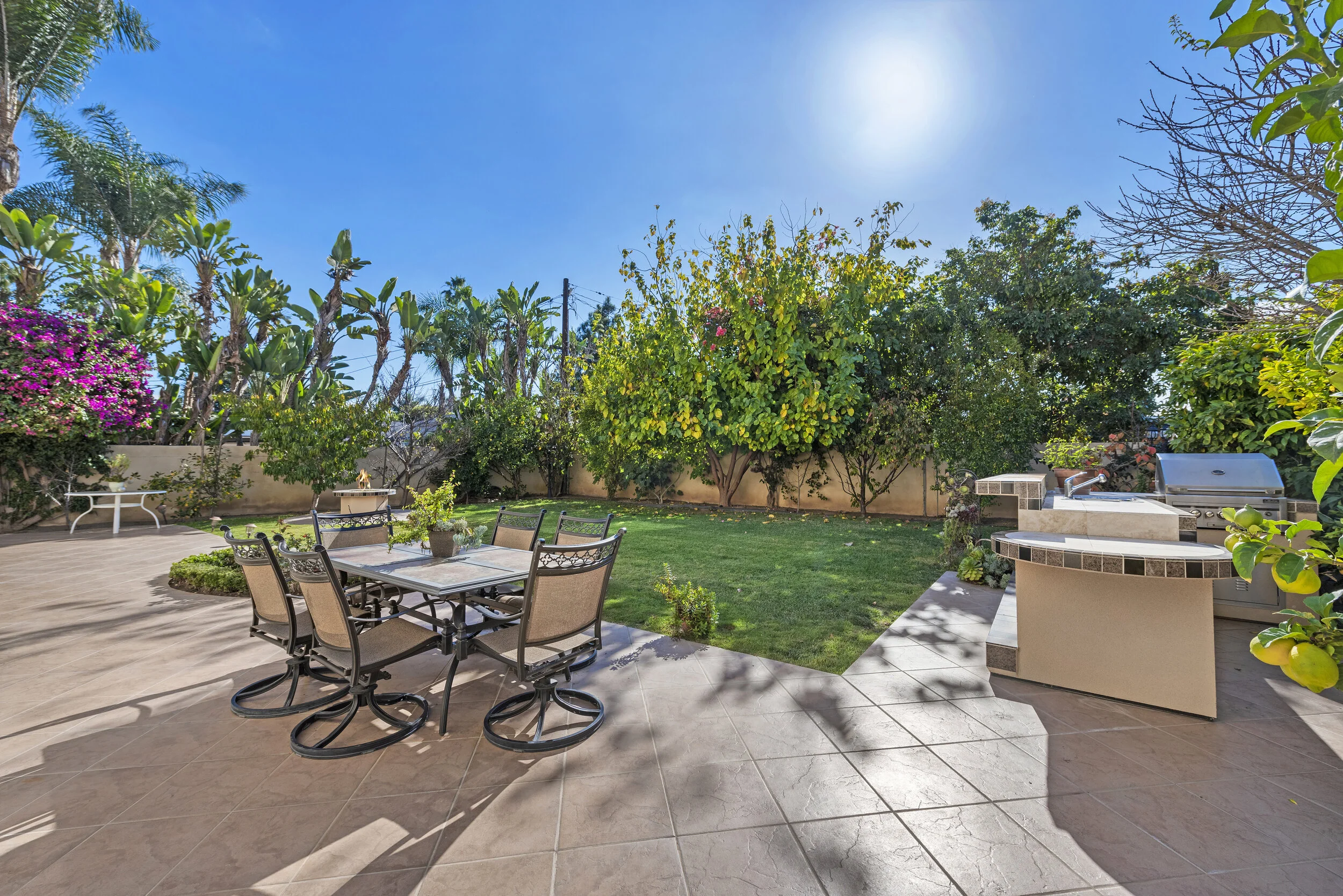 Backyard patio with outdoor dining table and six chairs, green lawn, trees, bushes, a built-in outdoor kitchen with a grill, sink, and counters, under a sunny blue sky.