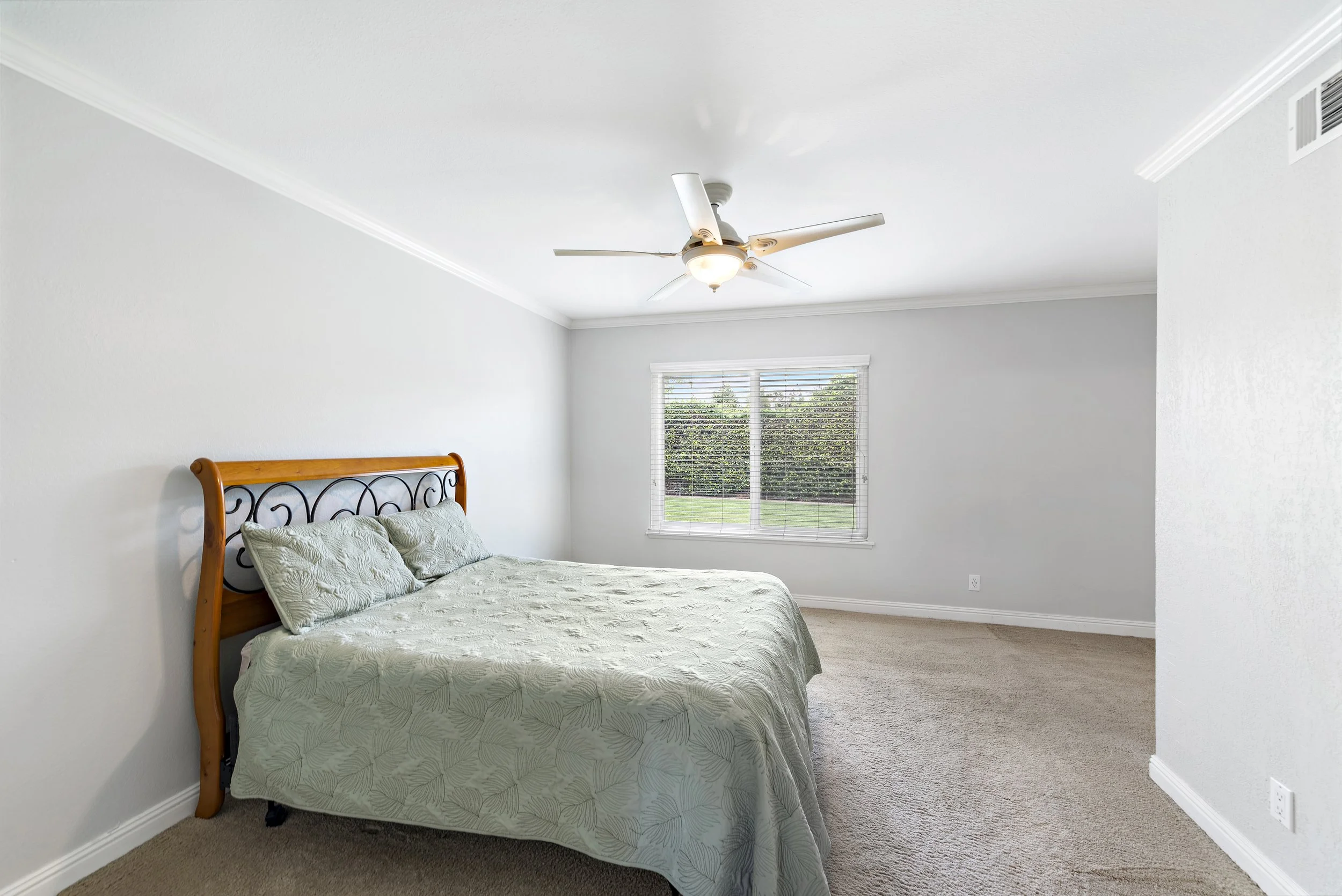 Empty bedroom with a bed, window, ceiling fan, white walls, beige carpet, and a view of green bushes outside.