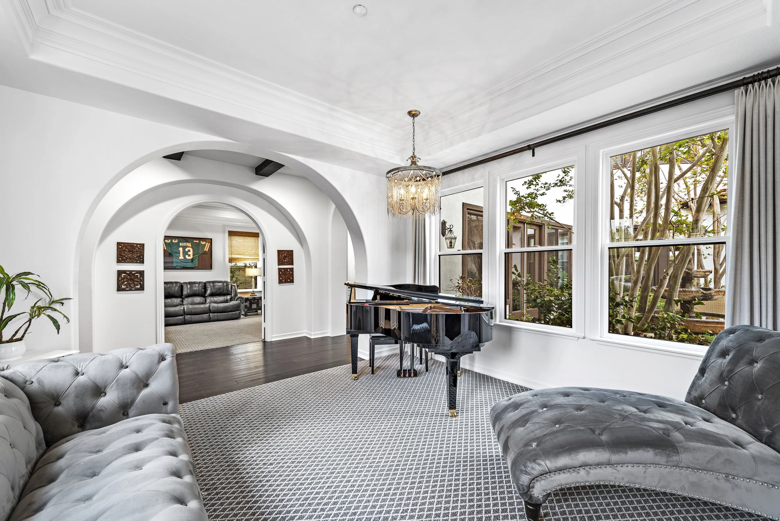 Living room with white walls, dark wood flooring, large windows, a black grand piano, gray tufted sofas, a chandelier, and a view into another room with a sports jersey framed on the wall.