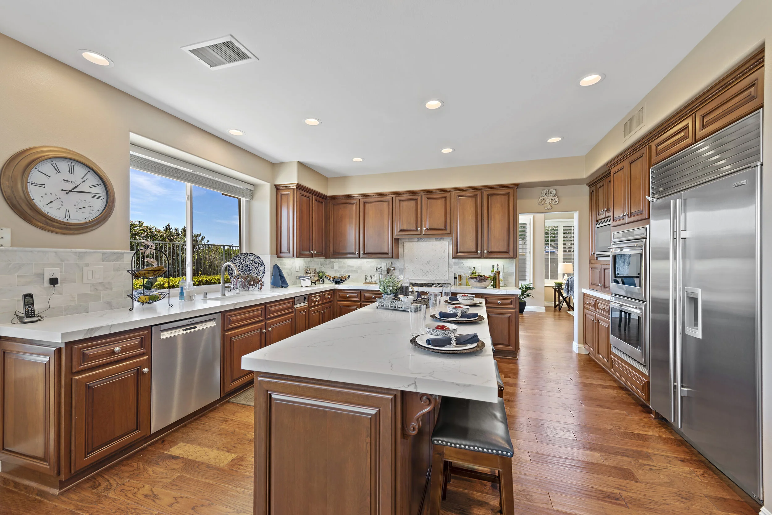 Modern kitchen with wooden cabinets, marble countertops, stainless steel appliances, and hardwood flooring, with a large window showing a sunny outdoor scene.