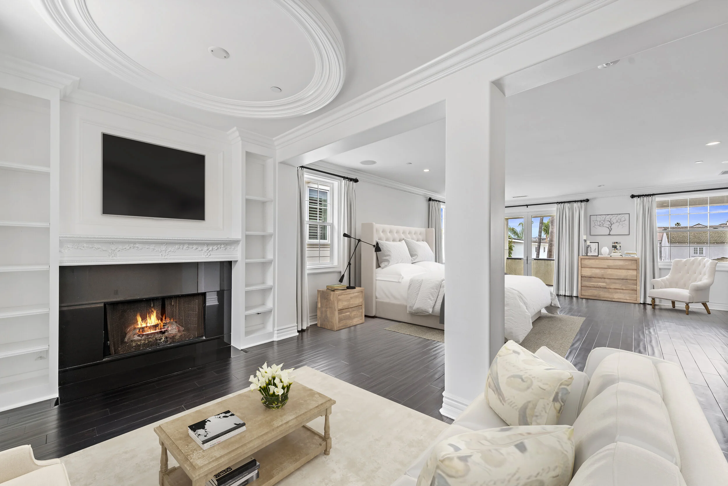 Bright, spacious bedroom with white walls, dark hardwood floors, a lit fireplace, a wall-mounted TV, and large windows with white curtains, featuring a bed with white bedding, a wooden nightstand, a beige armchair, and decorative elements.