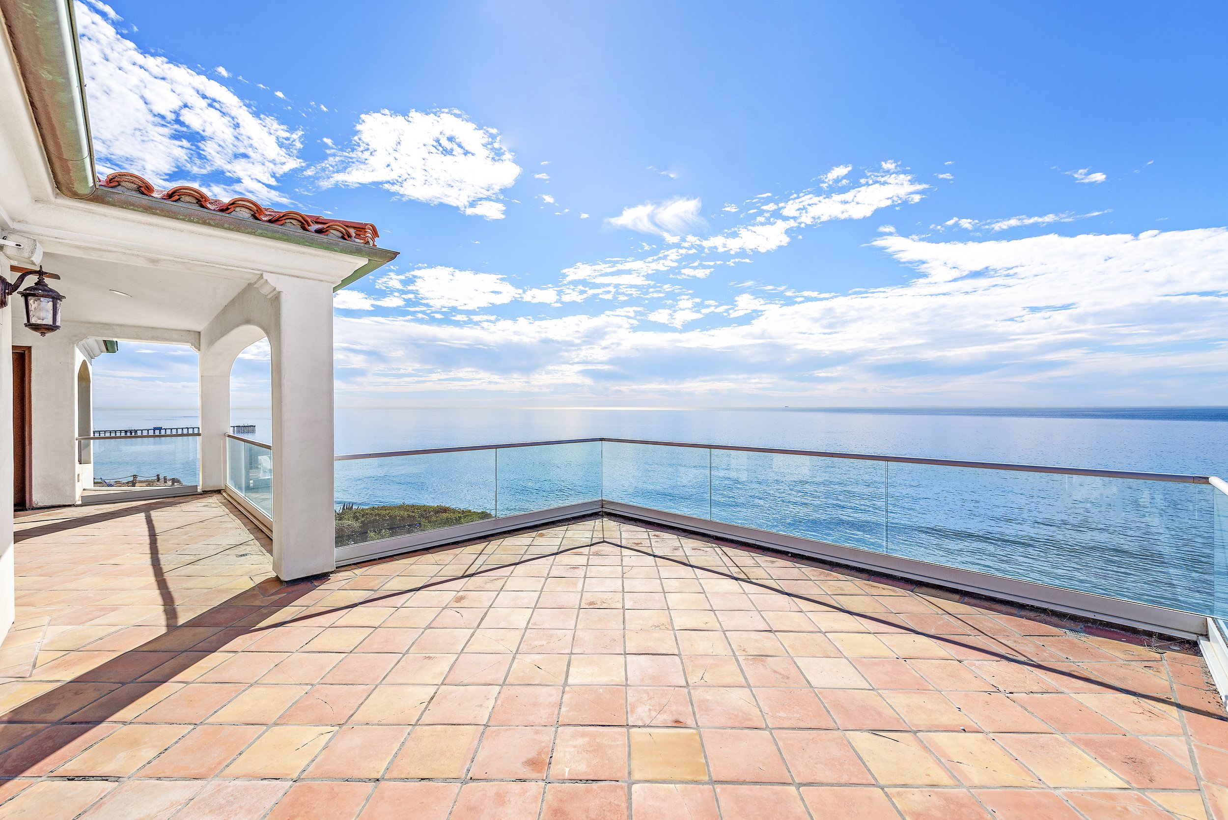 Empty terrace with terracotta tiles and glass railing overlooking the ocean under a partly cloudy blue sky.