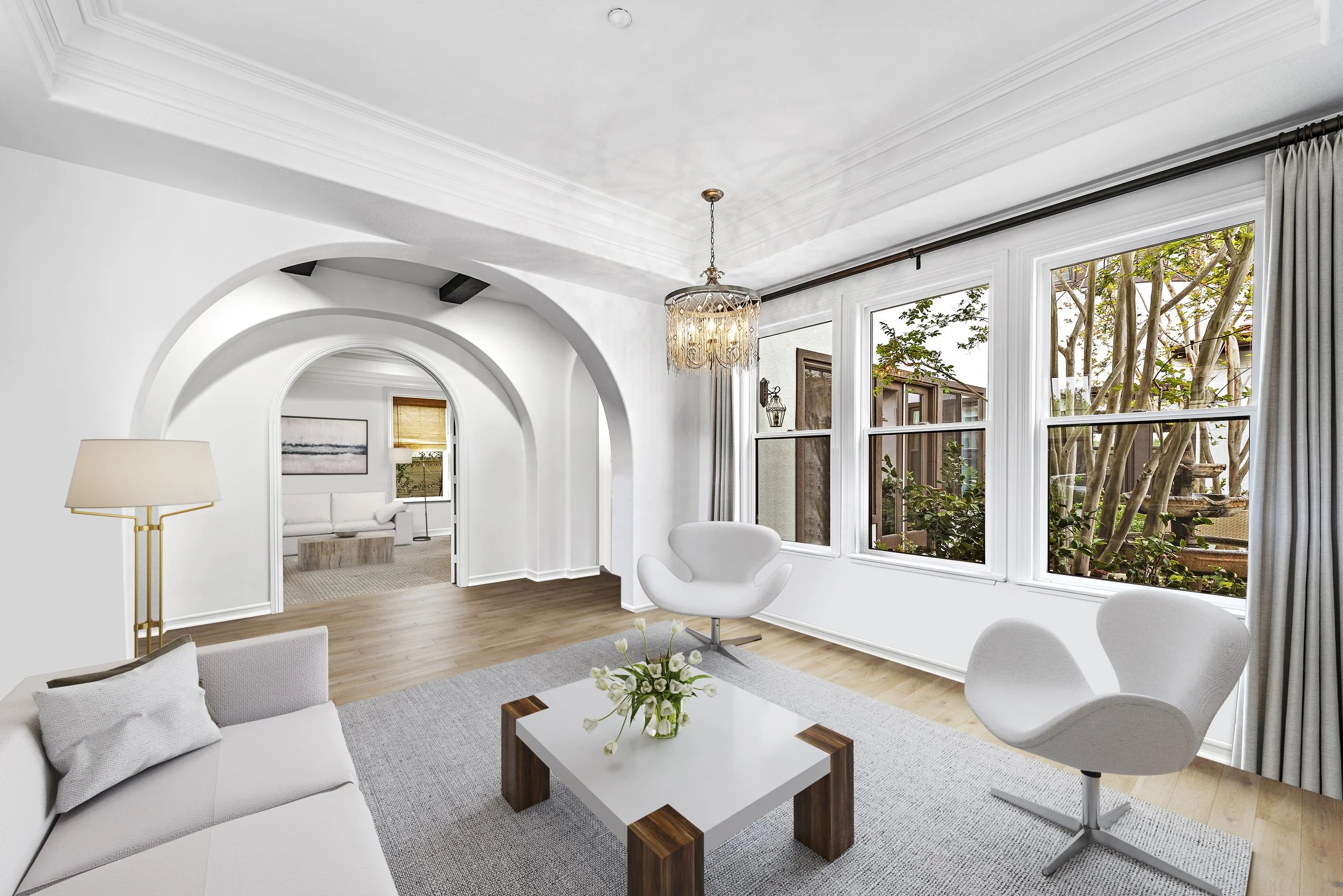 Bright living room with white walls, large windows, modern white chairs, a white sofa, a wooden coffee table with flowers, a hanging chandelier, and an arched doorway leading to another room.