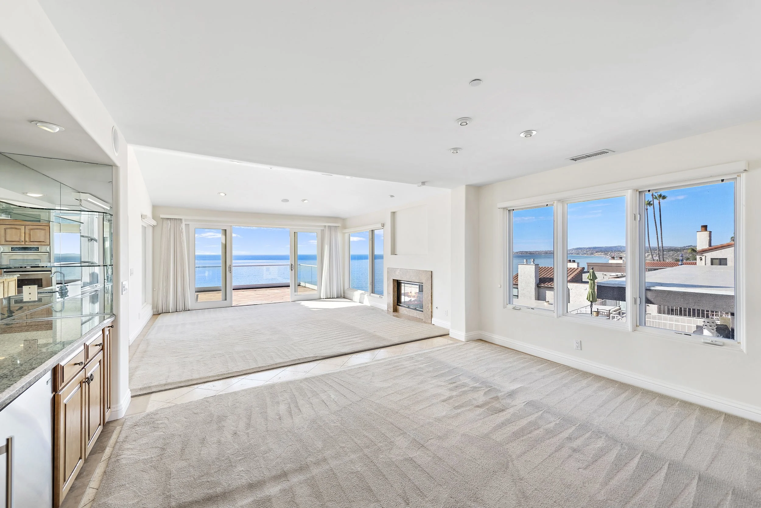 Bright living room with large windows and a view of the ocean, carpeted floor, fireplace, kitchen area on the left, and sliding glass doors leading to a balcony.