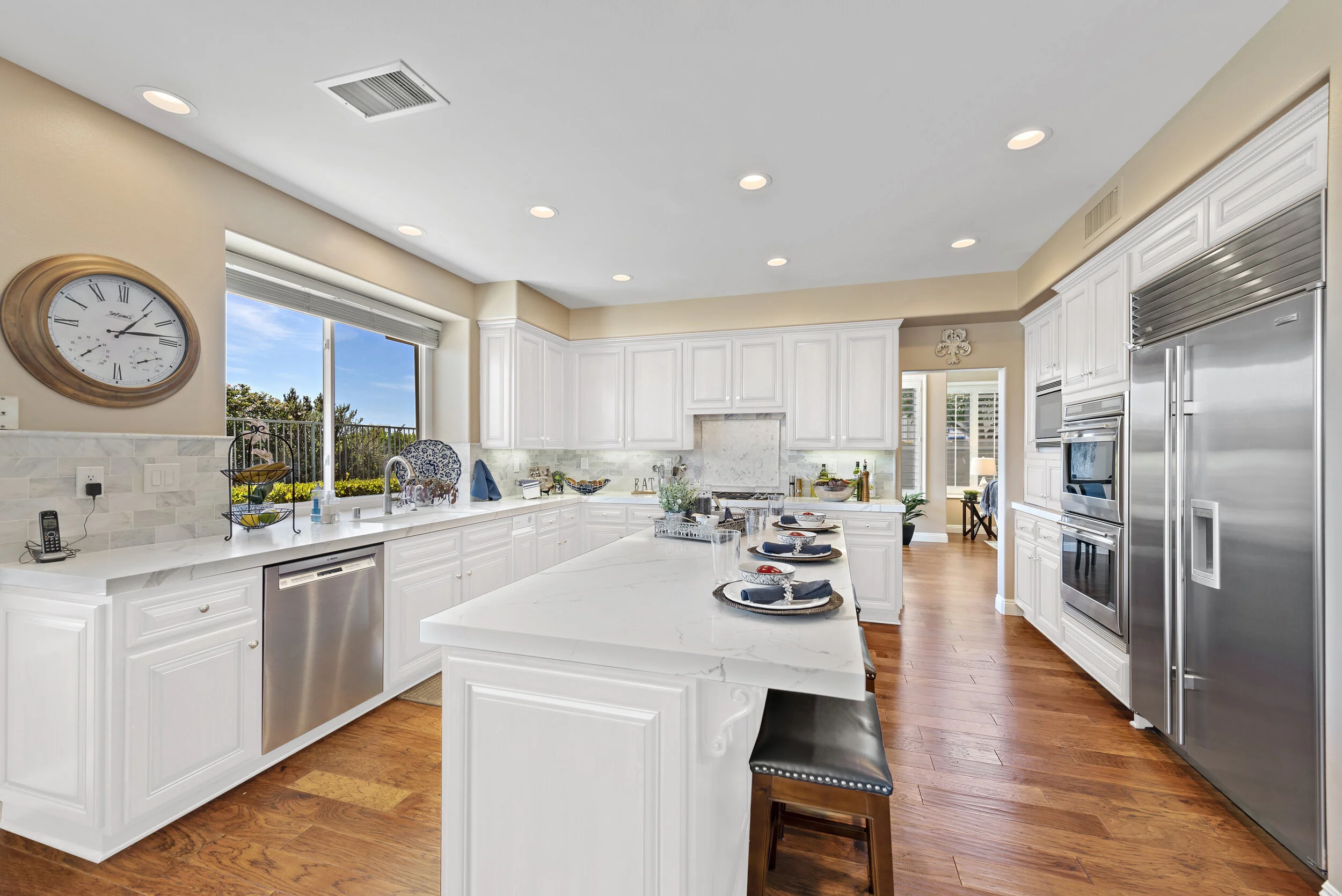 Bright kitchen with white cabinets, marble countertops, stainless steel appliances, hardwood floors, a large window, and a central island set with place settings.