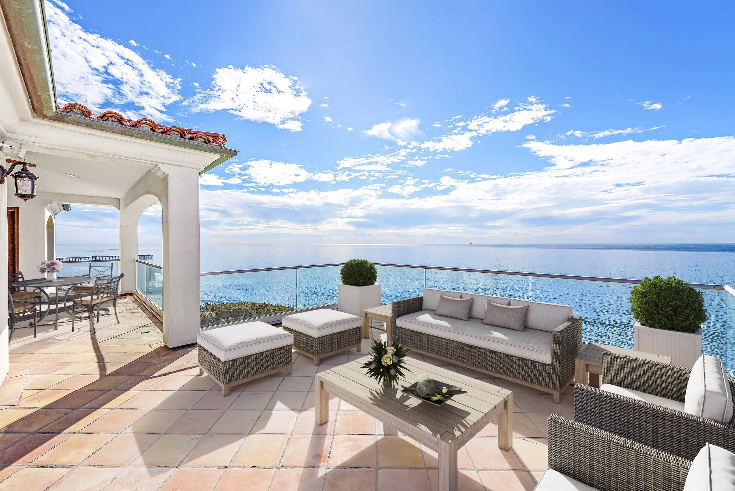 Oceanfront balcony with wicker and cushioned seating, potted plants, and a view of the blue sea and sky with clouds