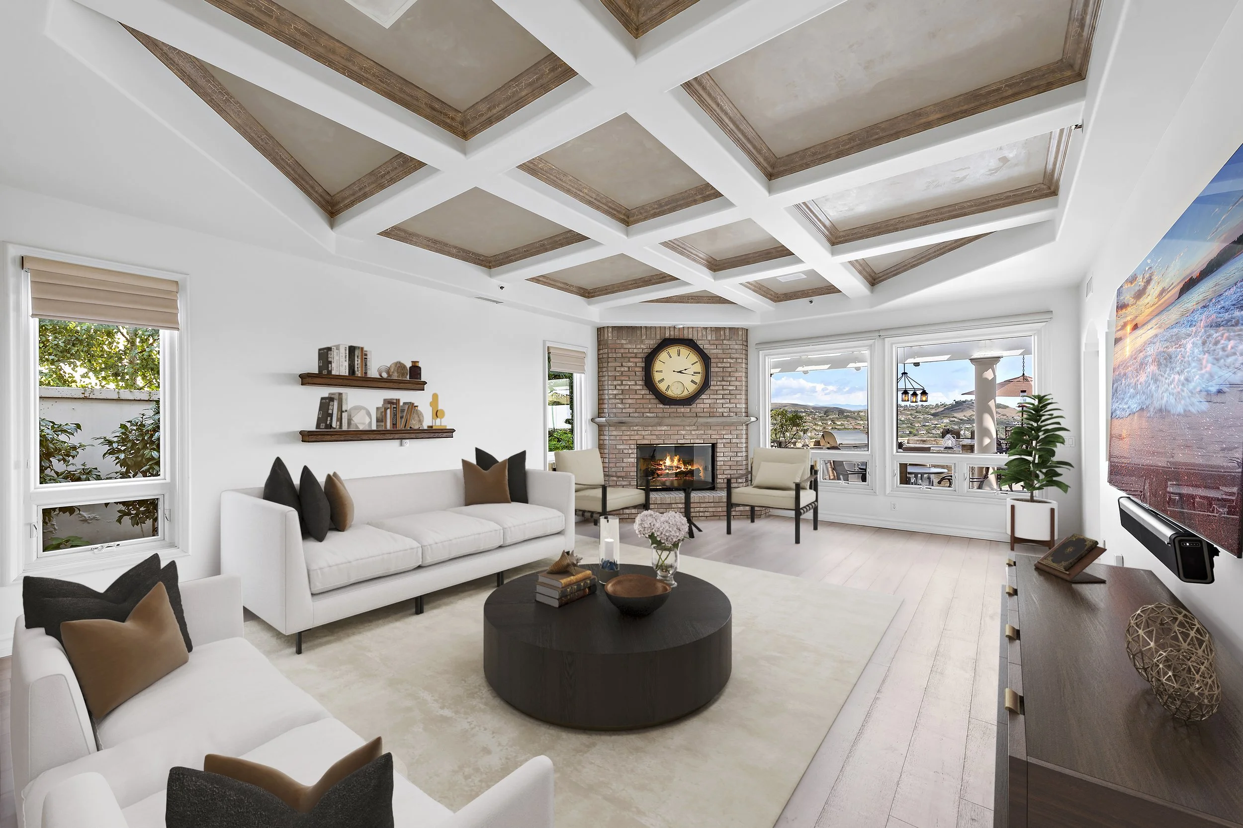 Bright living room with white walls, large windows, a brick fireplace with a clock, and modern furniture including a white sofa and armchairs. A large flat-screen TV is mounted on the wall, and a round dark wood coffee table holds decorative items. T