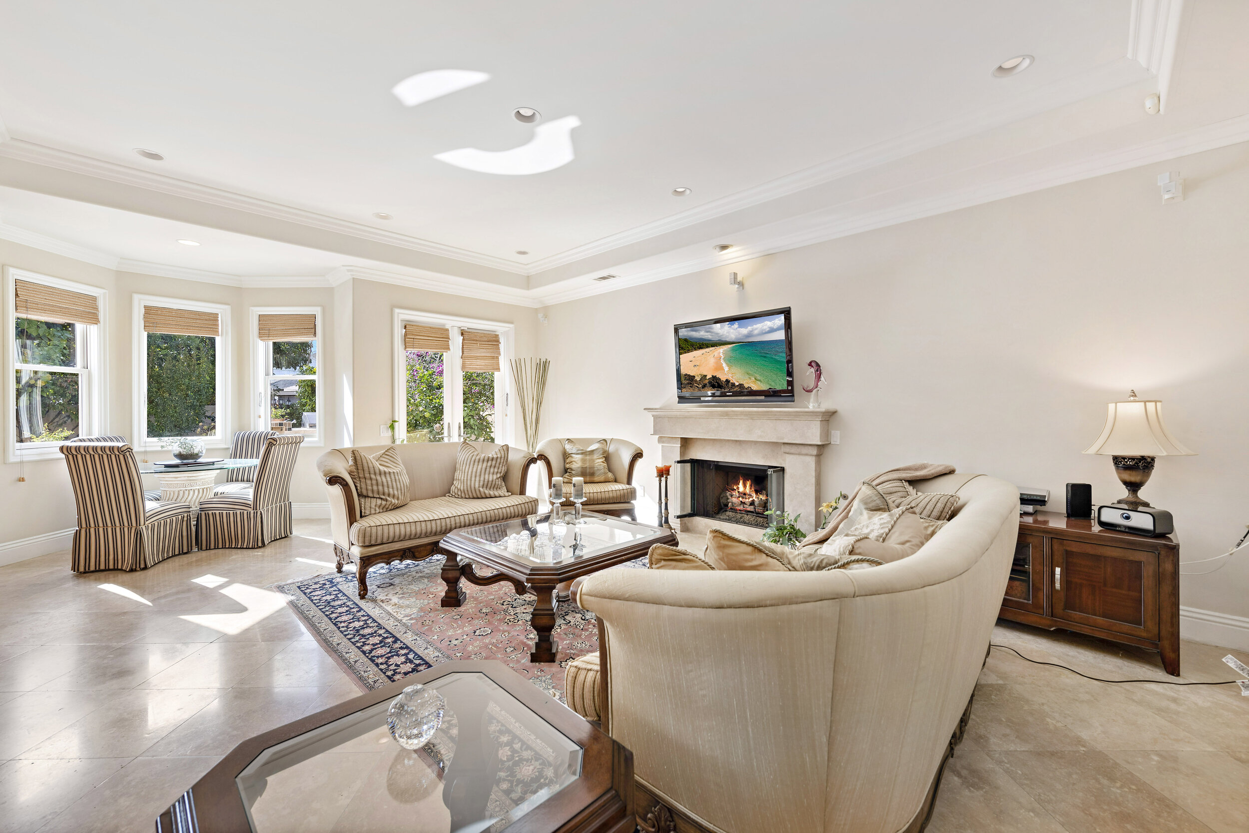 Bright living room with beige furniture, a fireplace, and a wall-mounted TV, with large windows and wooden blinds.