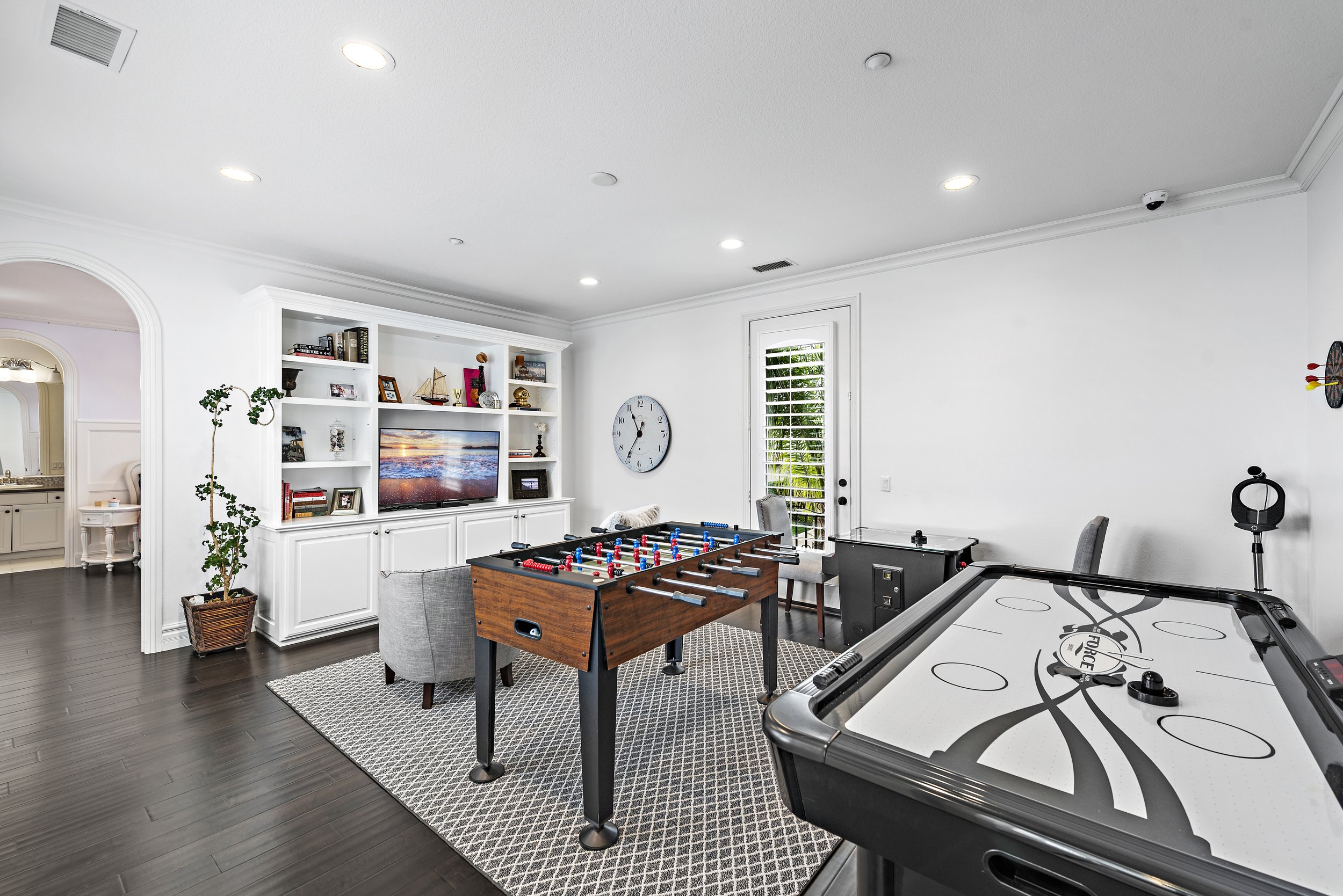 Game room with white walls and dark hardwood floors, featuring a foosball table, an air hockey table, a television on a white built-in entertainment center, and a clock on the wall next to a glass door with blinds, with a brick-patterned rug covering
