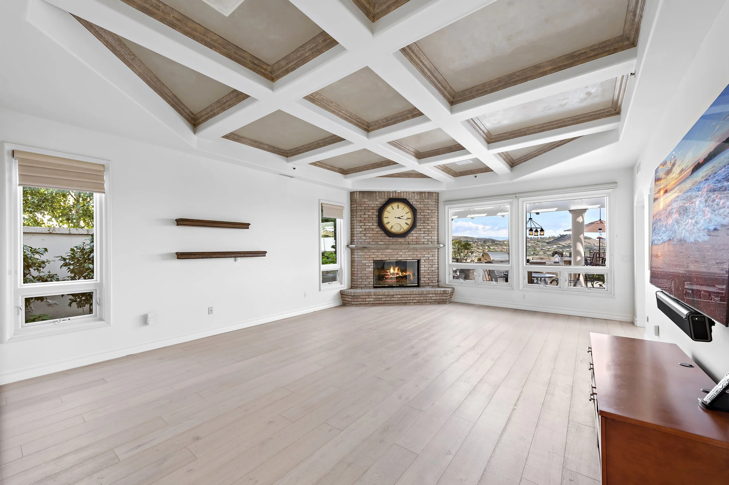Bright living room with large windows, fireplace, ceiling with wooden beams in a grid pattern, and a wall-mounted TV.