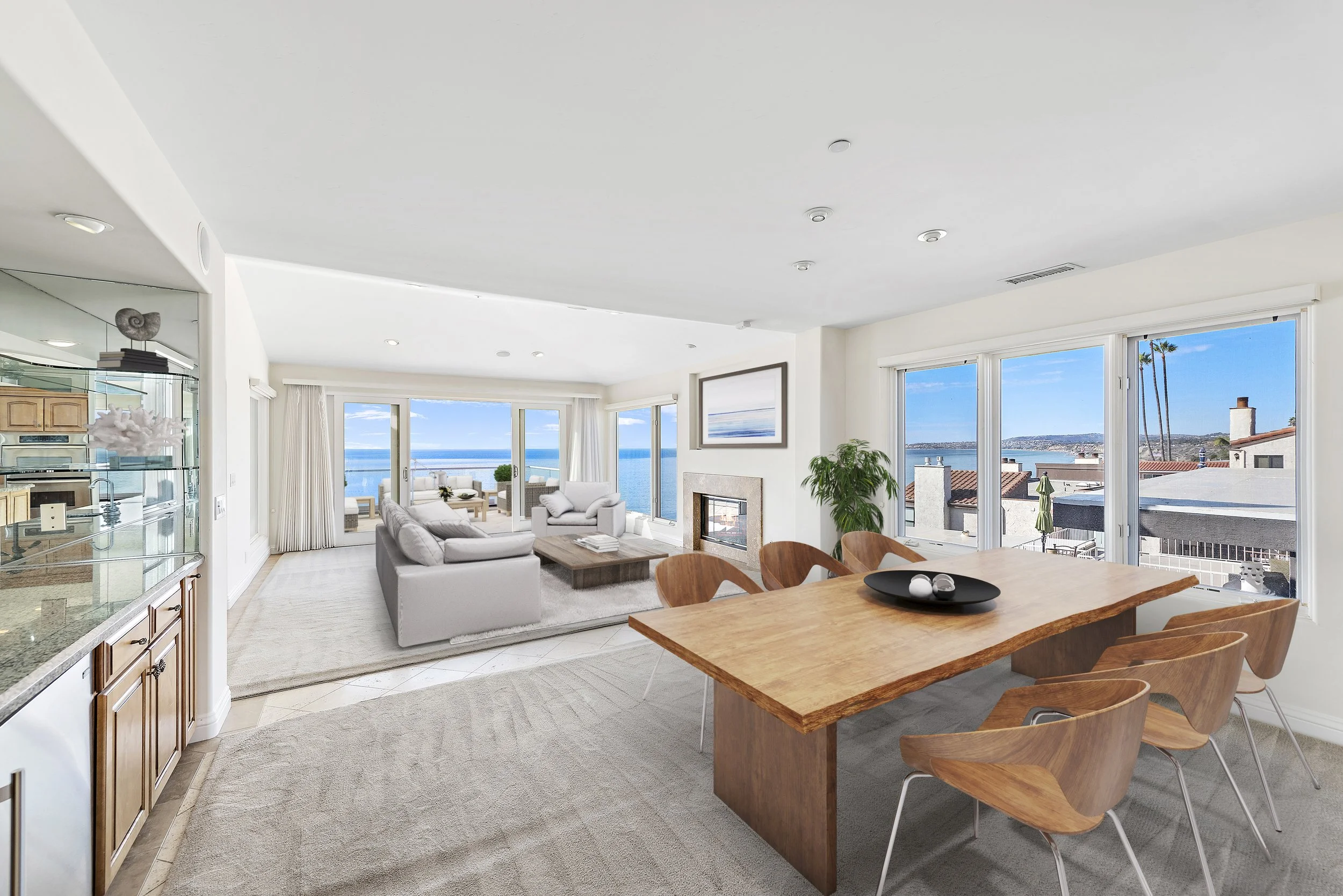 Bright, modern living and dining room with large windows and ocean view, furnished with white sofas, wooden dining table with six chairs, fireplace, and indoor plants.