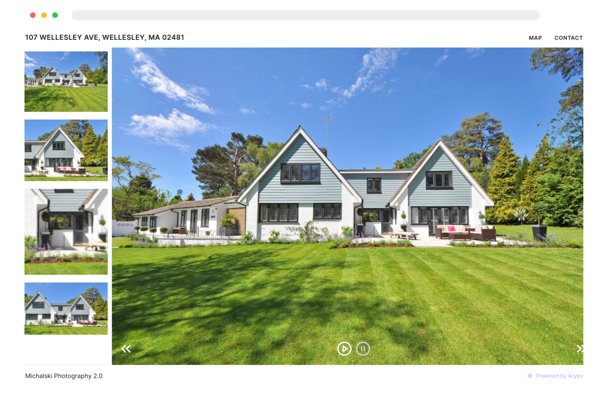 Medium-sized house with multiple gabled roofs, gray siding, black window frames, and front porch with outdoor furniture on a bright, sunny day. Surrounding lush green lawn and trees under a blue sky with some clouds.