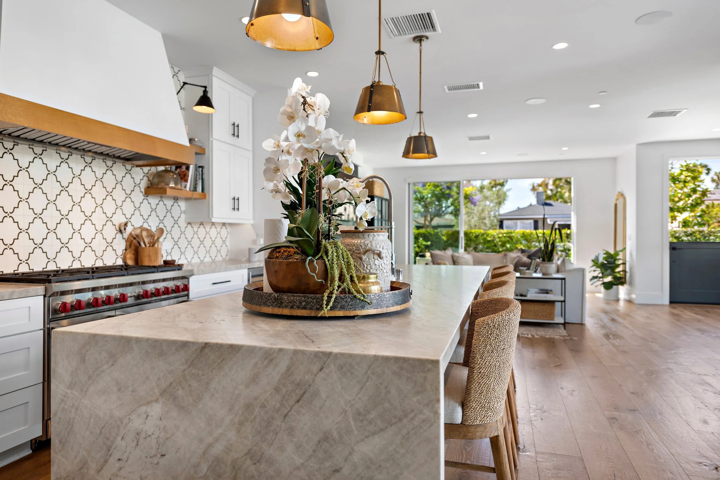 Modern open-plan kitchen and living room with a marble island, pendant lighting, and large windows showing greenery outside.
