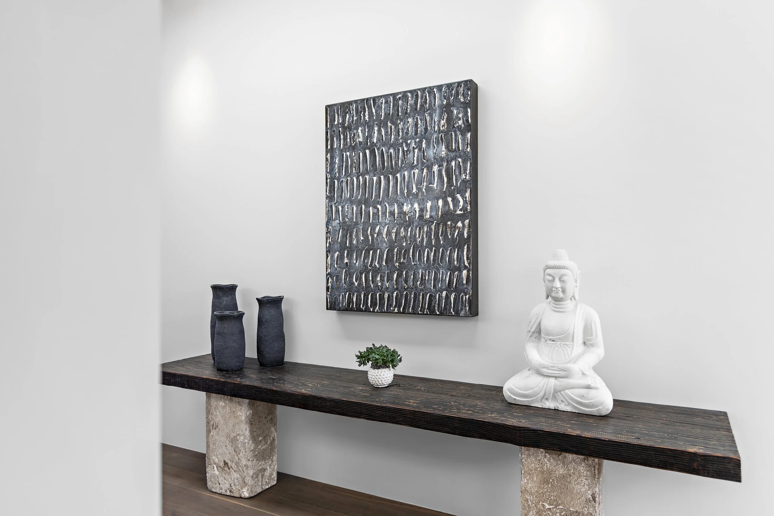 Minimalist interior decor with a dark wooden table supported by two stone legs, a white Buddha statue, three tall black vases, and a small potted plant, with a textured artwork on a white wall.