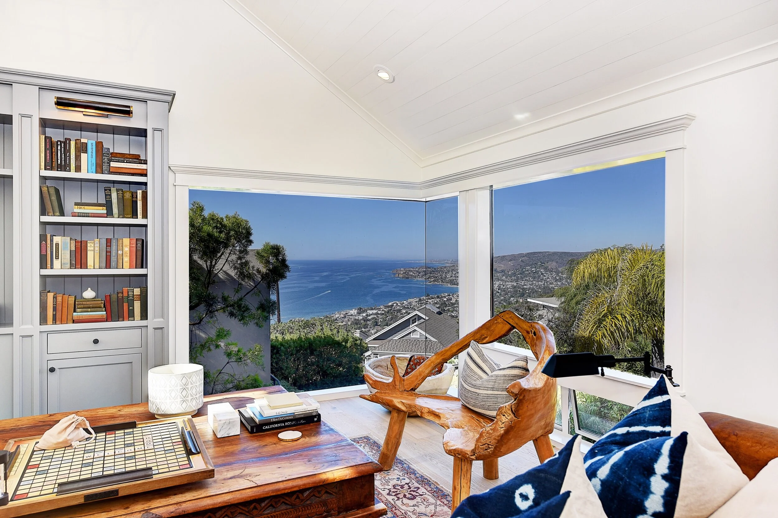 Room with large windows showing a coastal view, a wooden desk with books and a game board, a wooden chair with a striped pillow, and a bookshelf filled with books.