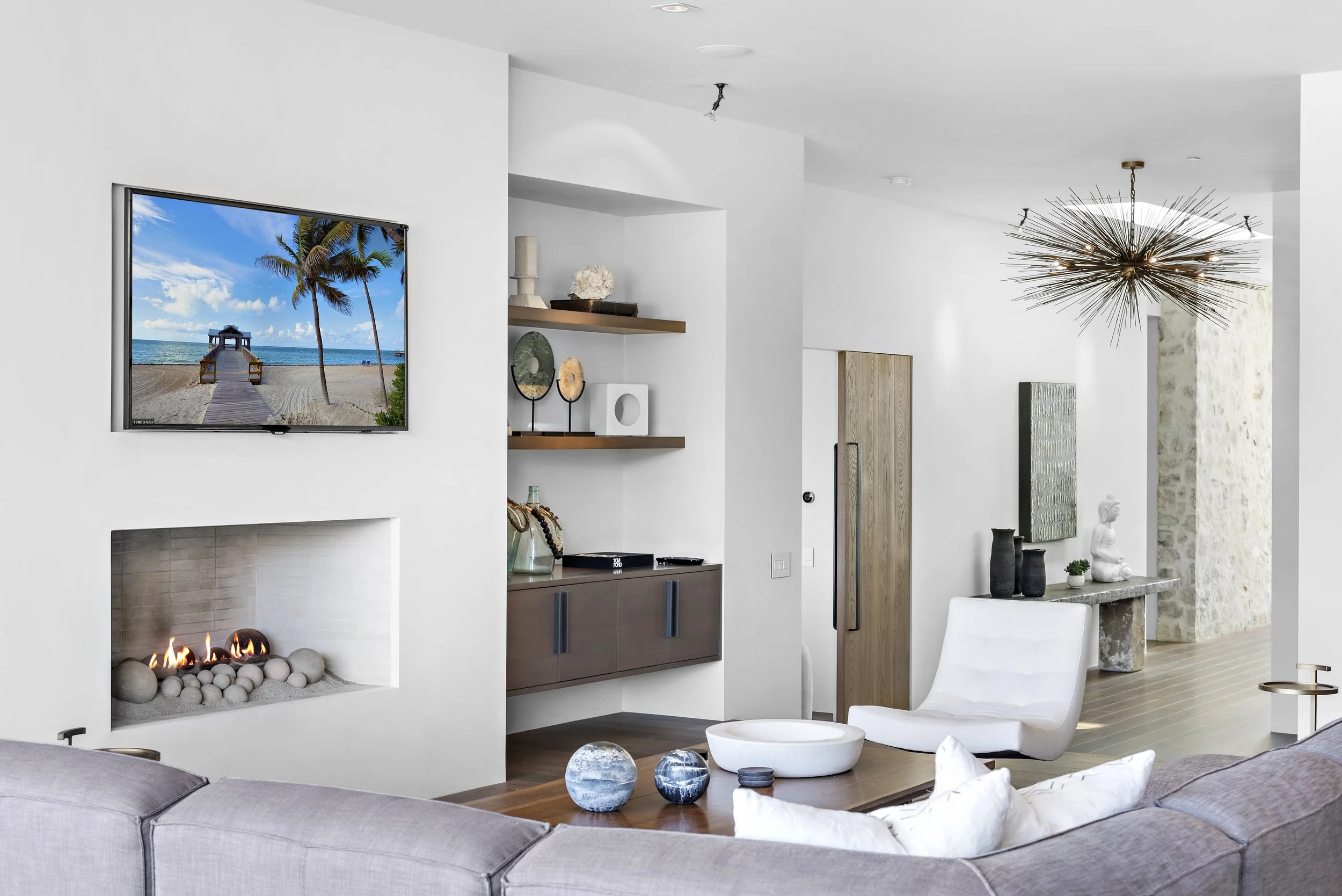 Modern living room with a built-in fireplace, wall-mounted flat-screen TV displaying a beach scene, white walls, wooden flooring, a white lounge chair, decorative vases, sculptures, and a unique starburst chandelier.