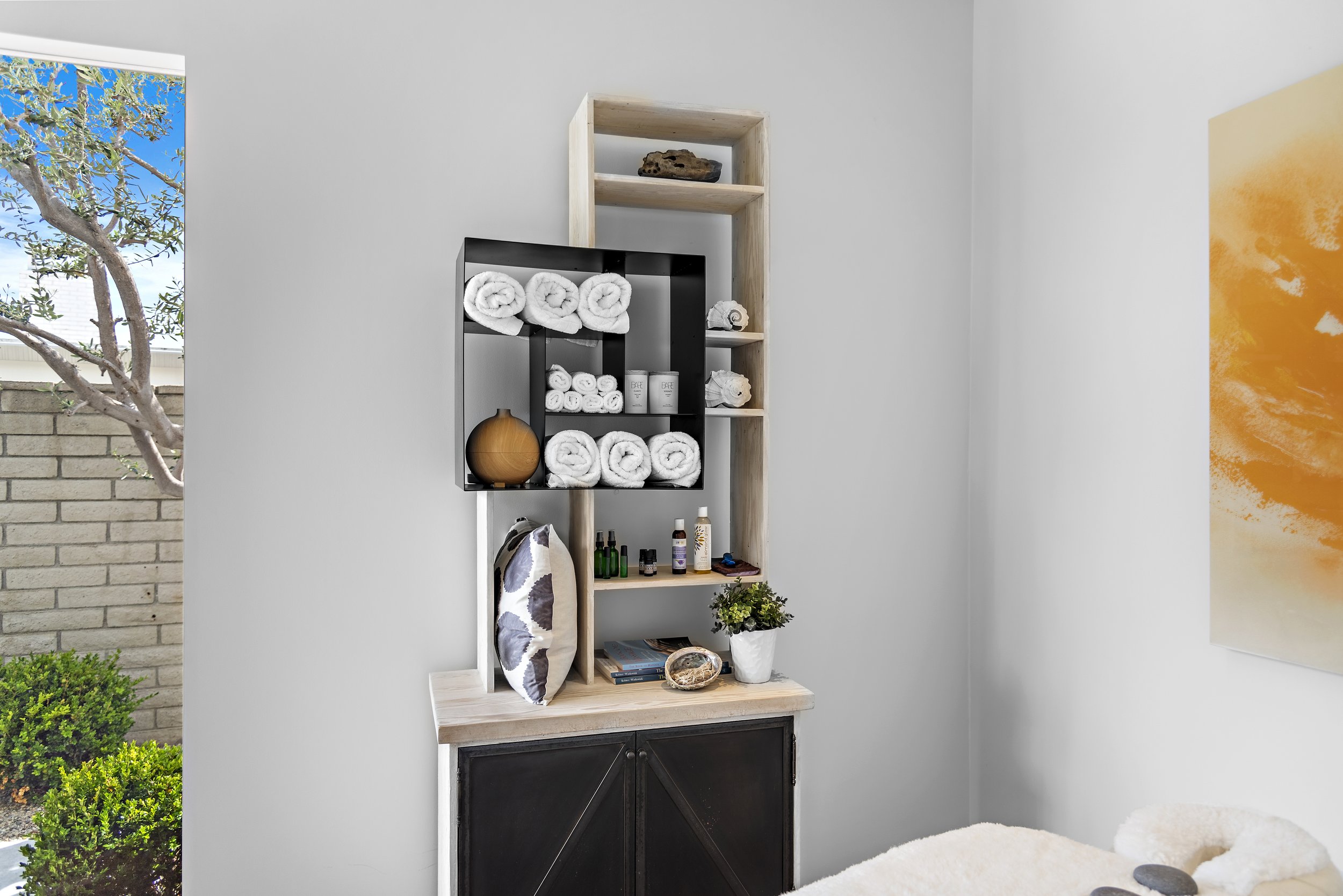 Bathroom shelf with neatly rolled white towels, skincare products, and small bottles, next to a window and a framed artwork with orange and yellow hues.