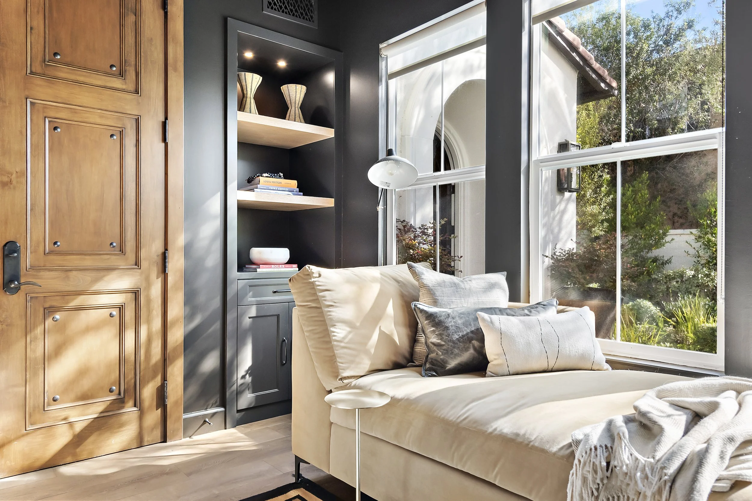 Sunlit living room with large window, beige sofa with multiple pillows, wooden door, and built-in dark gray shelving with decorative objects and books.