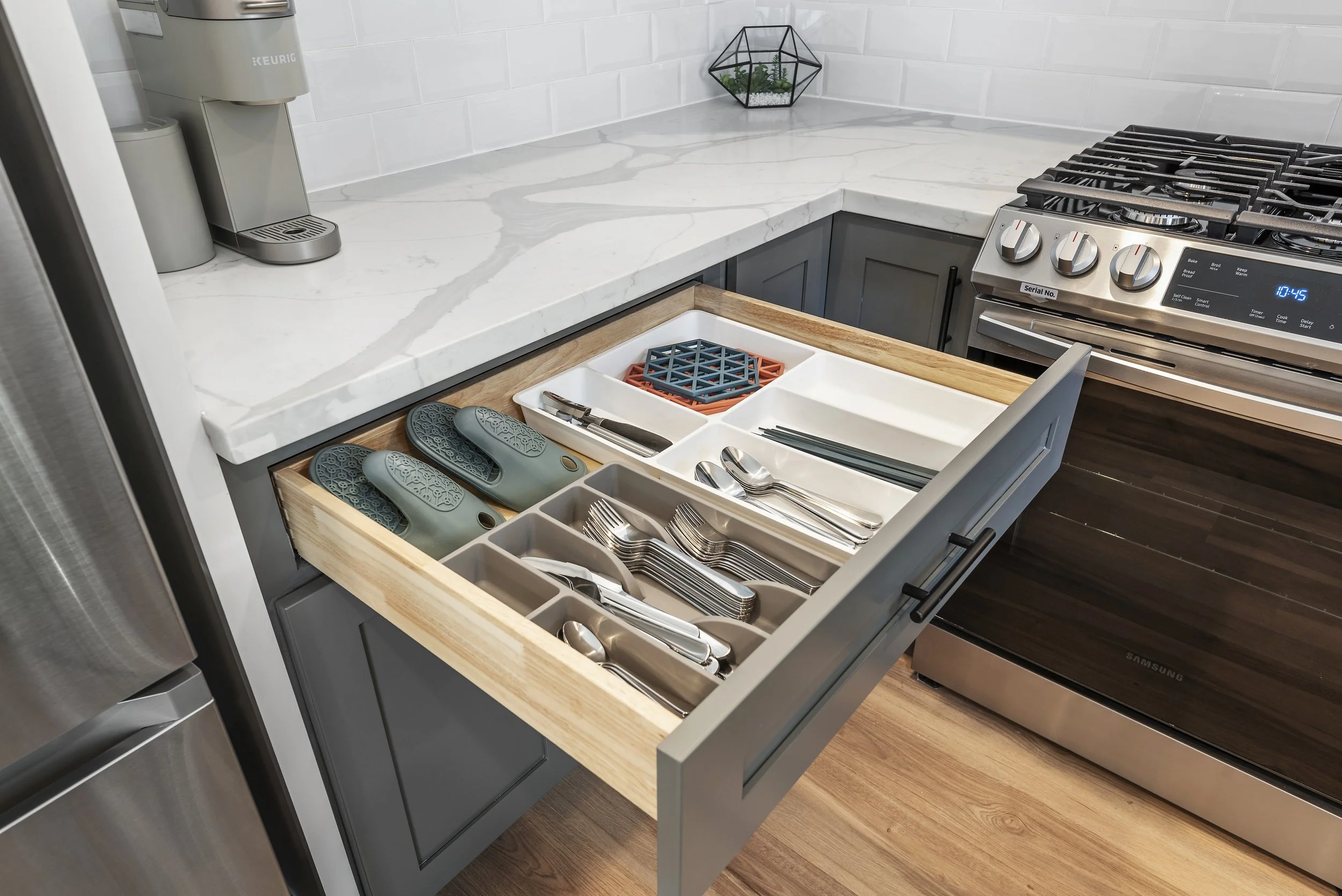 Open kitchen drawer with organized silverware, utensils, and rubber slippers, next to a marble countertop and a stove.