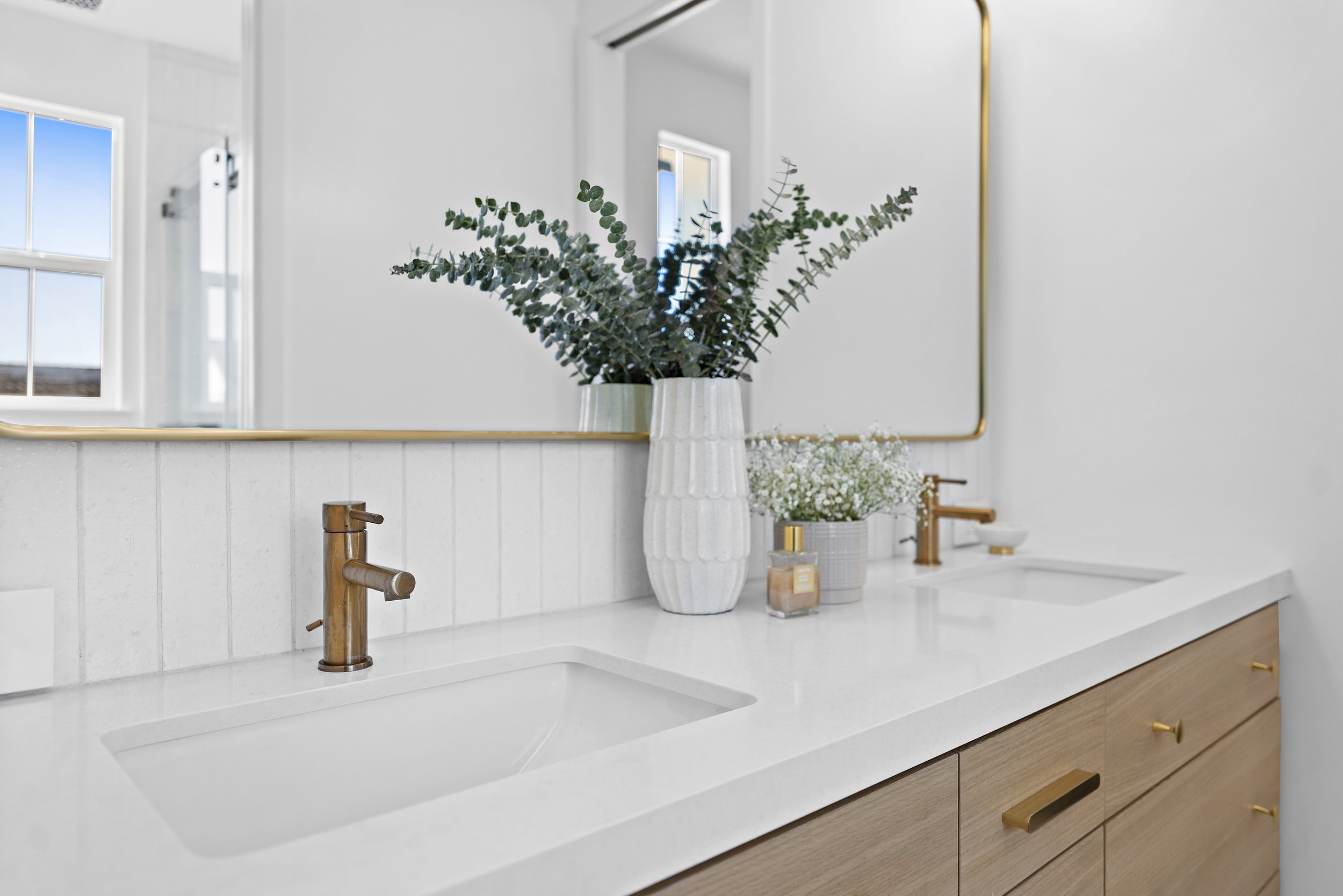 Bathroom vanity with a large mirror, white countertop, two sinks, gold faucets, a large white vase with green foliage, a smaller vase with white flowers, a decorative soap dispenser, and a second gold faucet at the far end.