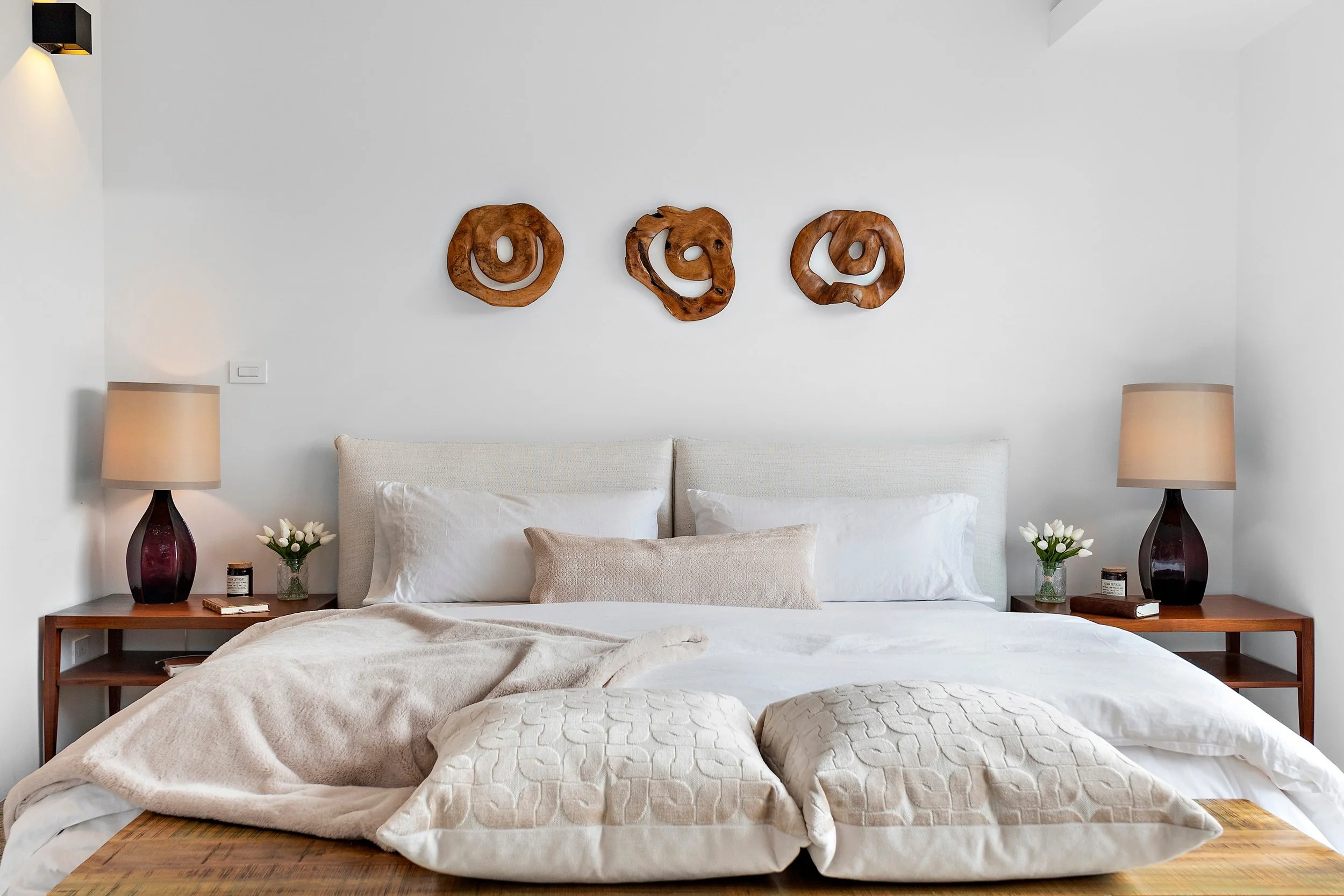 A neatly made bed with white linens, beige pillows, and a beige blanket, flanked by two wooden nightstands with matching lamps and small vases of white flowers, in a bright, minimalist bedroom with white walls decorated with three abstract wooden wal