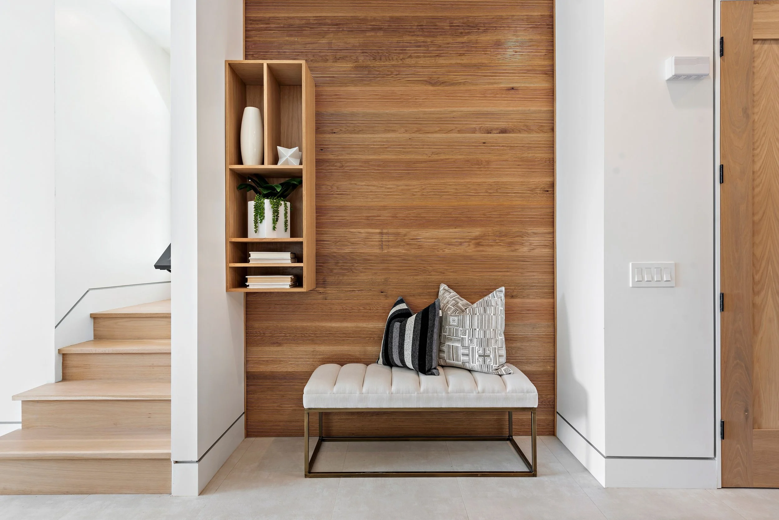 Modern entryway with wooden staircase, wall-mounted wooden shelf with decorative items and plant, beige bench with patterned pillows, white and wooden walls, and a vent on the right wall.