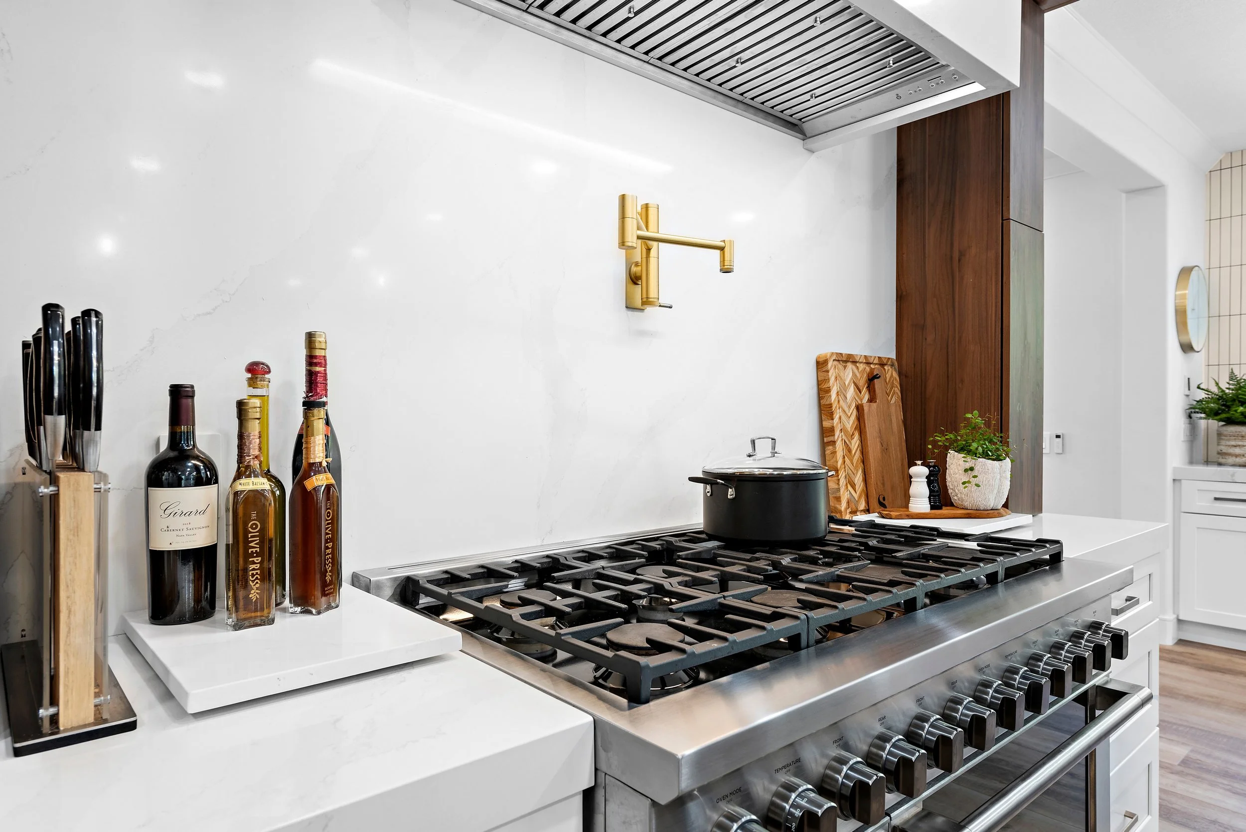 Modern kitchen with a stainless steel stove, black pot, wooden cutting boards, bottles of wine and olive oil, knife set, salt and pepper shakers, plant, and drawers with wood and white finishes.