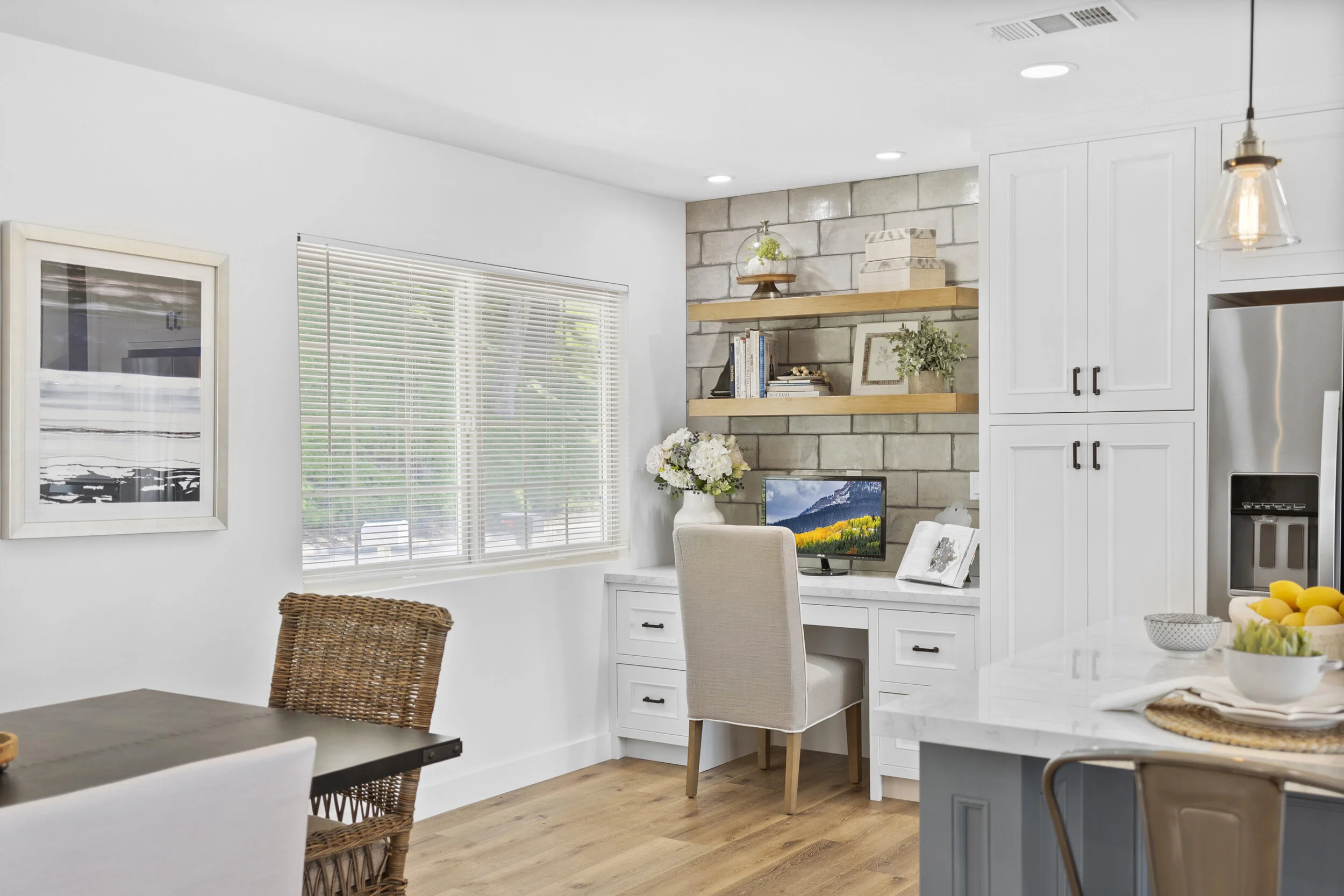 A cozy, modern kitchen corner with a small workspace, featuring white cabinetry, a desk with a computer monitor, beige upholstered chair, floating wooden shelves with decor and books, window with blinds, a vase of flowers, and a brick accent wall.
