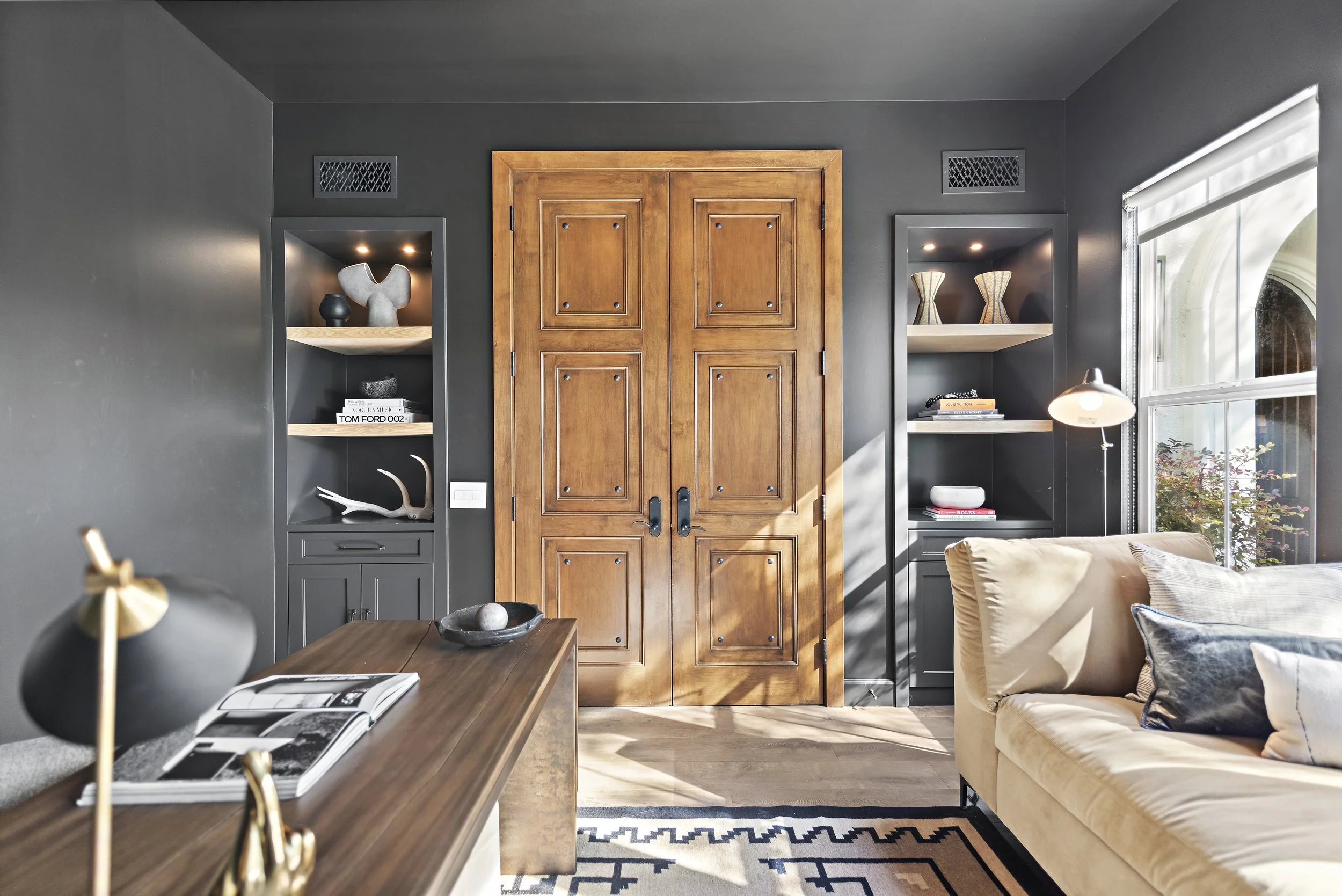 Living room with dark gray walls, wooden double doors, built-in shelves with decor, a beige sofa with pillows, a wooden side table, and a window with sunlight streaming in.
