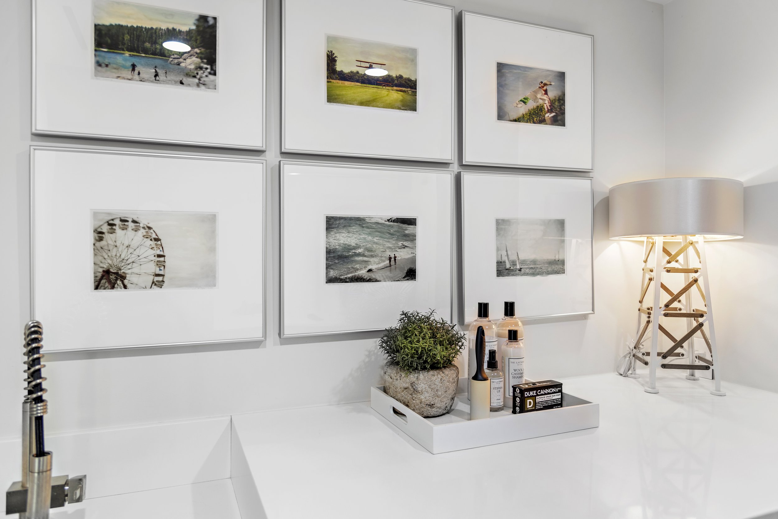 Living room corner with six framed photos on white wall, a lamp with a wooden structure, a white tray with skincare products and a potted plant, and a faucet visible on the left.
