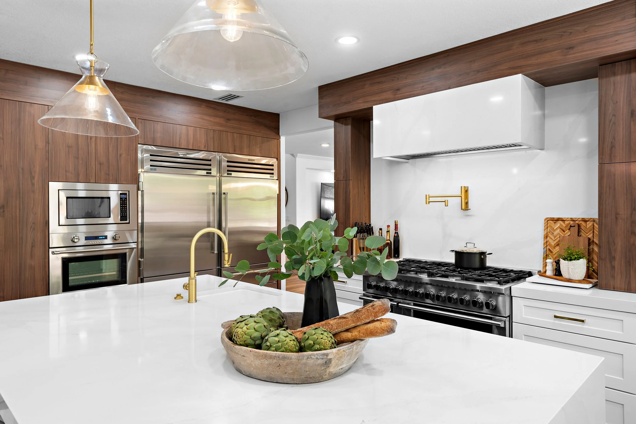 Modern kitchen with white island counter, gold faucet, artichokes and bread in a bowl, wooden cabinets, stainless steel refrigerator, built-in microwave and oven, black stovetop with pot, decorative items, and a potted plant.